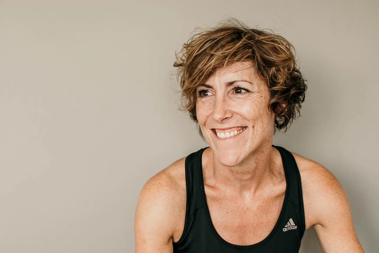 A woman with short, curly brown hair wearing a black Adidas tank top, smiling and looking to her right, against a plain light gray background.