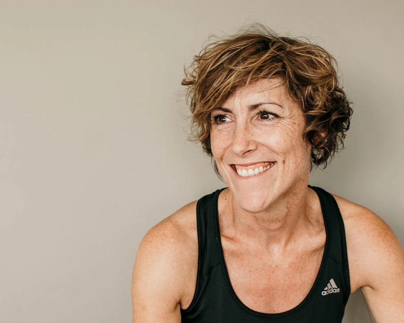A smiling woman with short, curly brown hair and freckles wears a black Adidas sports top, looking to the side against a plain light background.