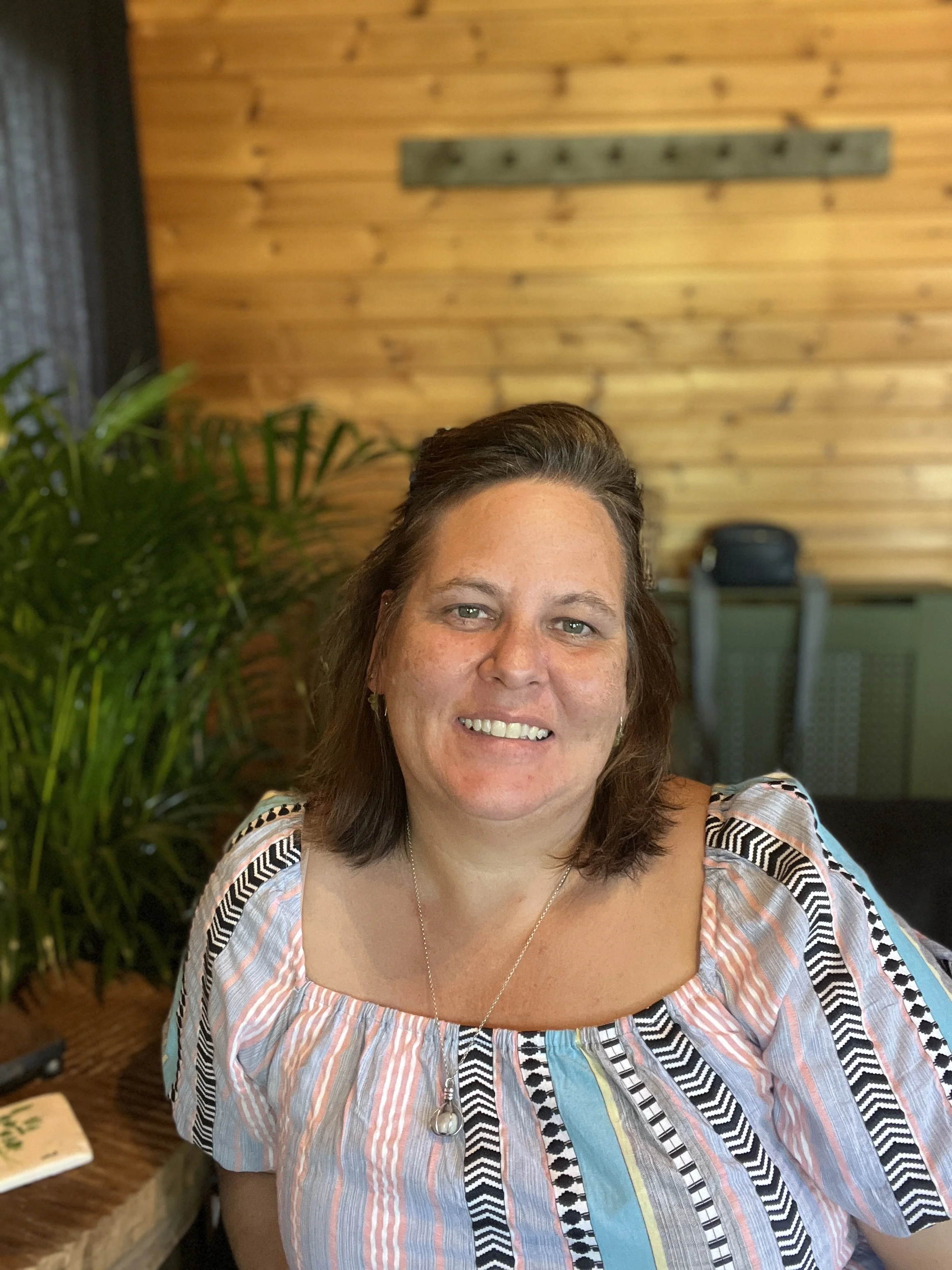 A woman smiling at the camera inside a room with wooden walls and green plants.