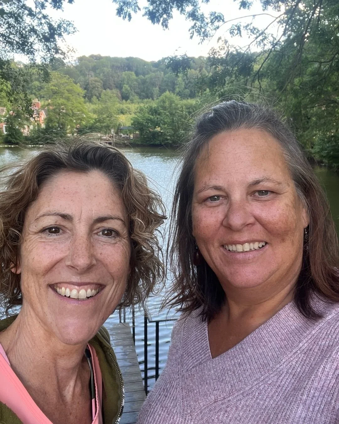 2 midlife women standing in front of a lake. One with wavy mid brown hair the other with darker shoulder length hair wearing a pink jumper
