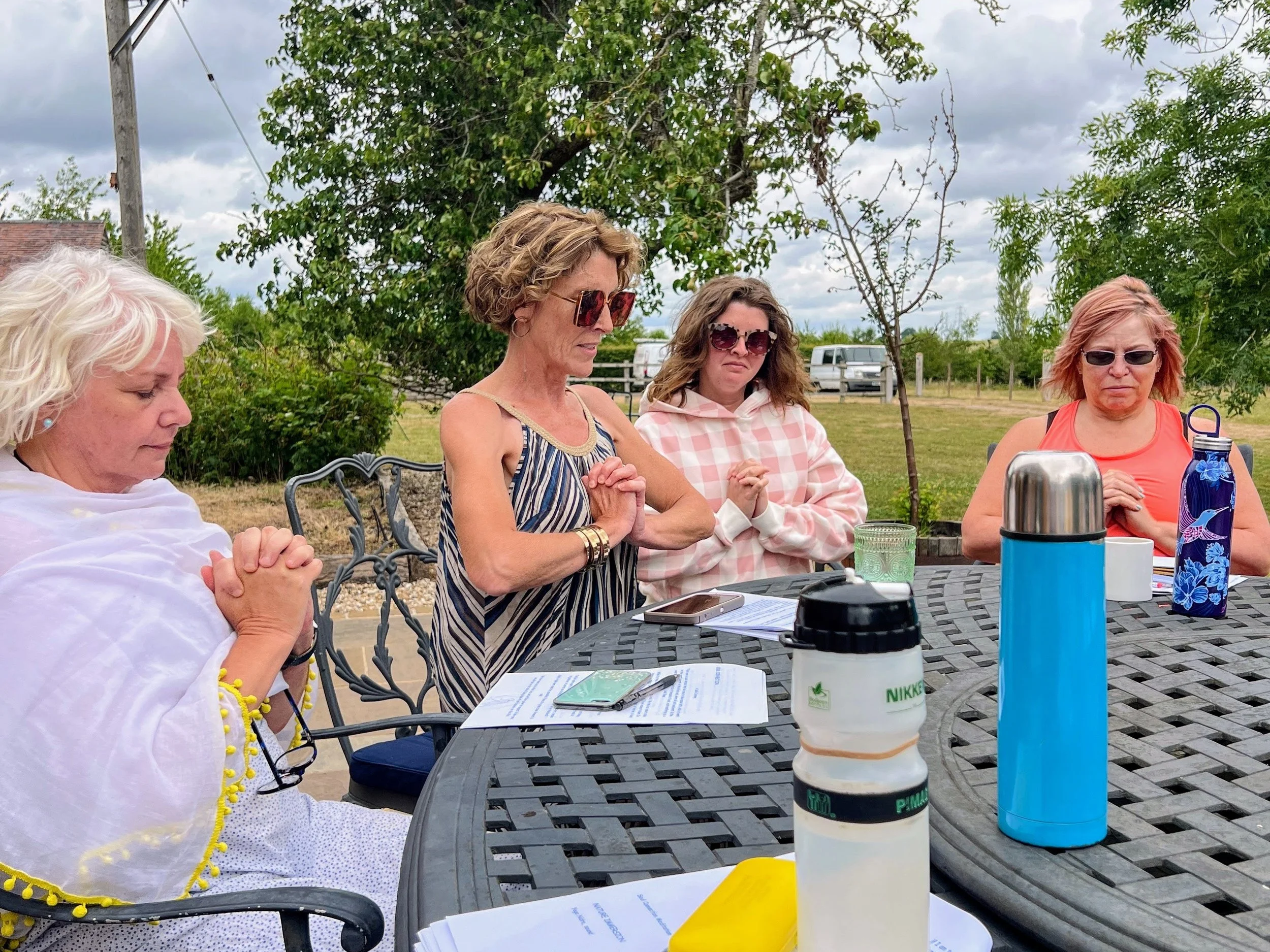 Four women sitting at an outdoor table praying with hands clasped meditating, papers, and water bottles on the table, in a backyard with trees and cloudy sky.