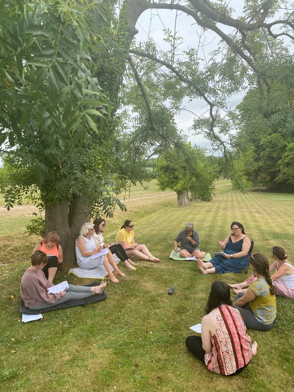 A group of people sitting outside on the grass under a large tree, participating in a discussion or workshop, with some holding notebooks and papers.