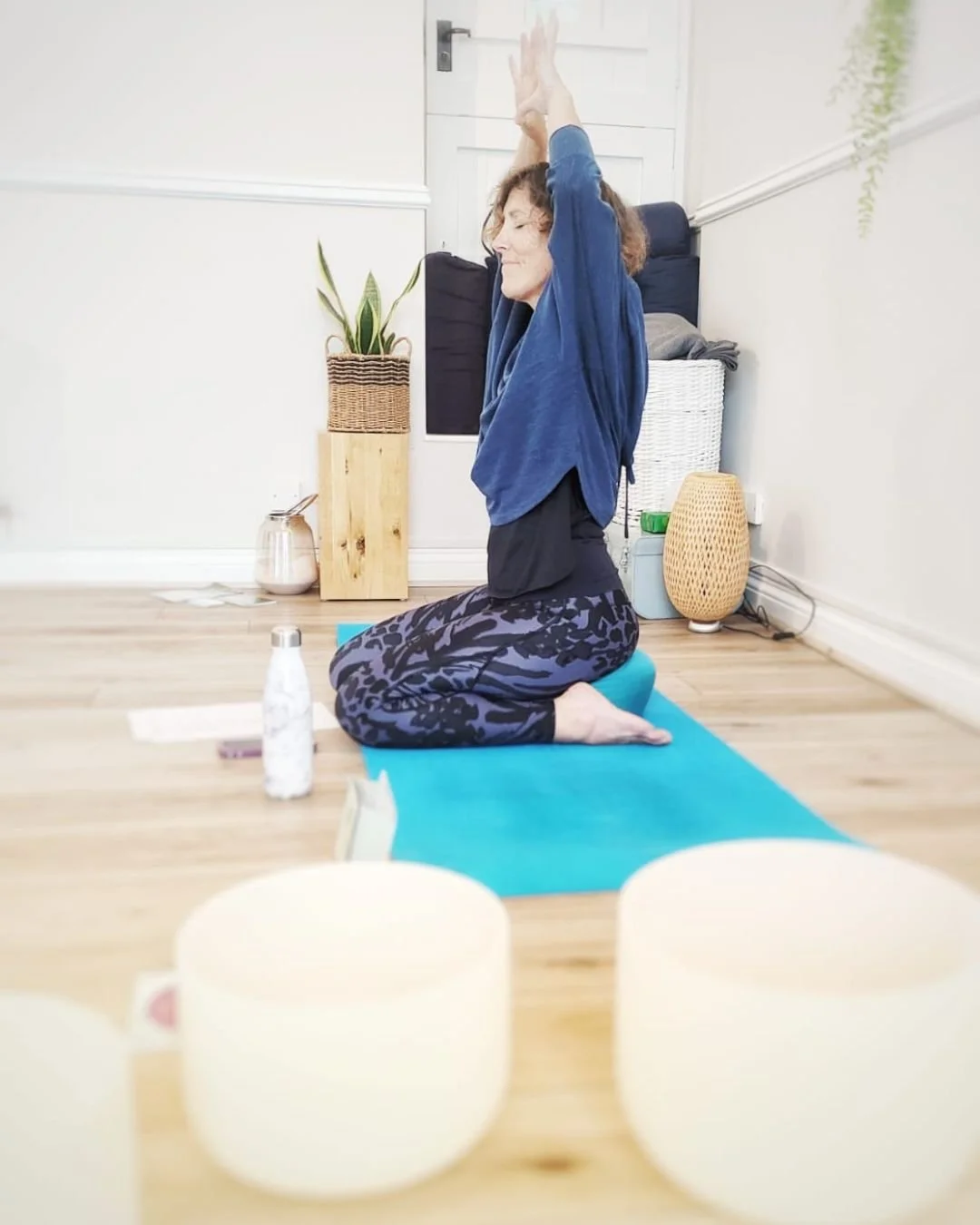 Alex  practicing yoga kneeling on a blue mat with arms raised overhead in a room with white walls, wooden floor, and decorative plants.