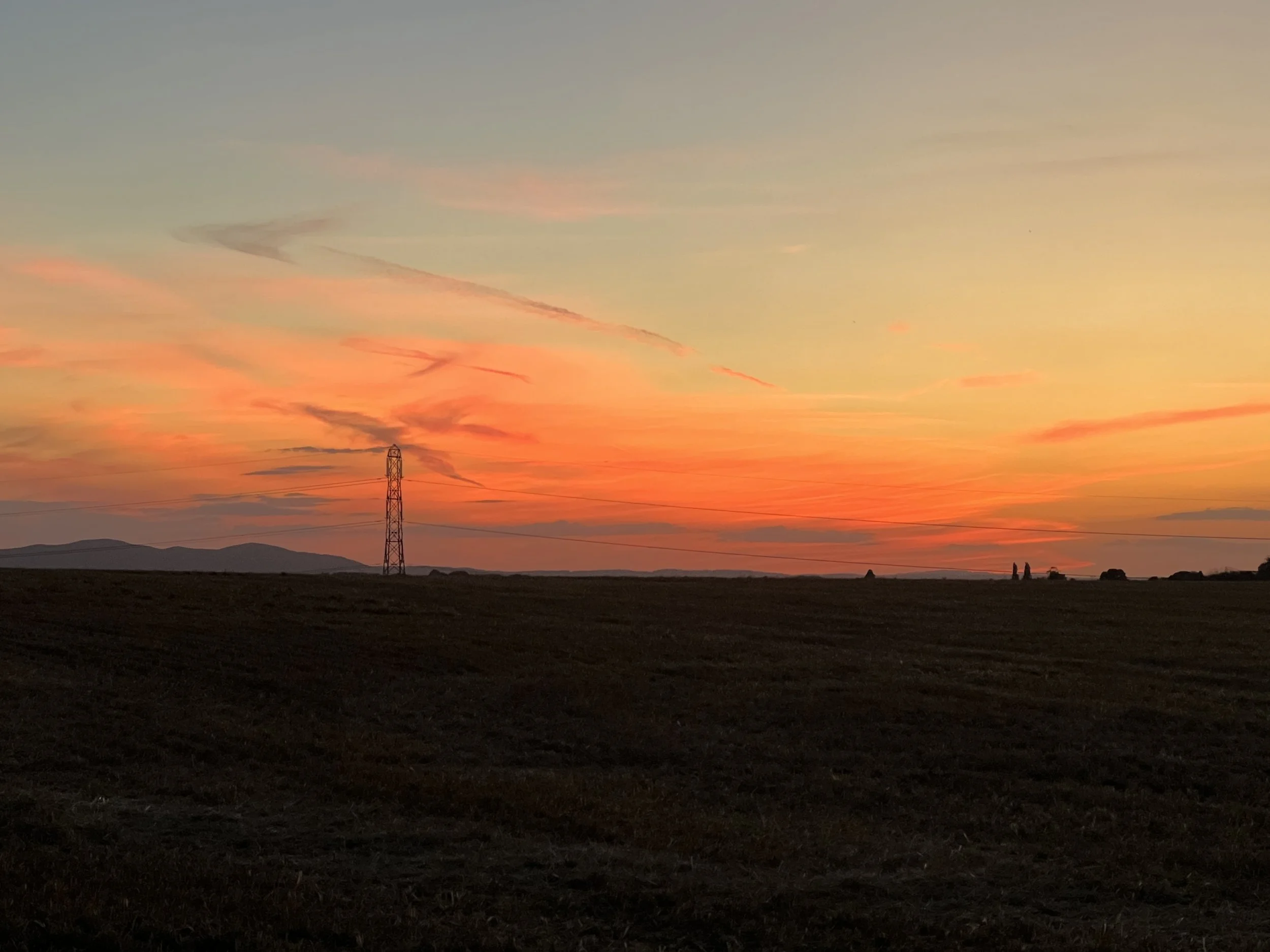 Sunset over a rural landscape with a power line and distant hills.