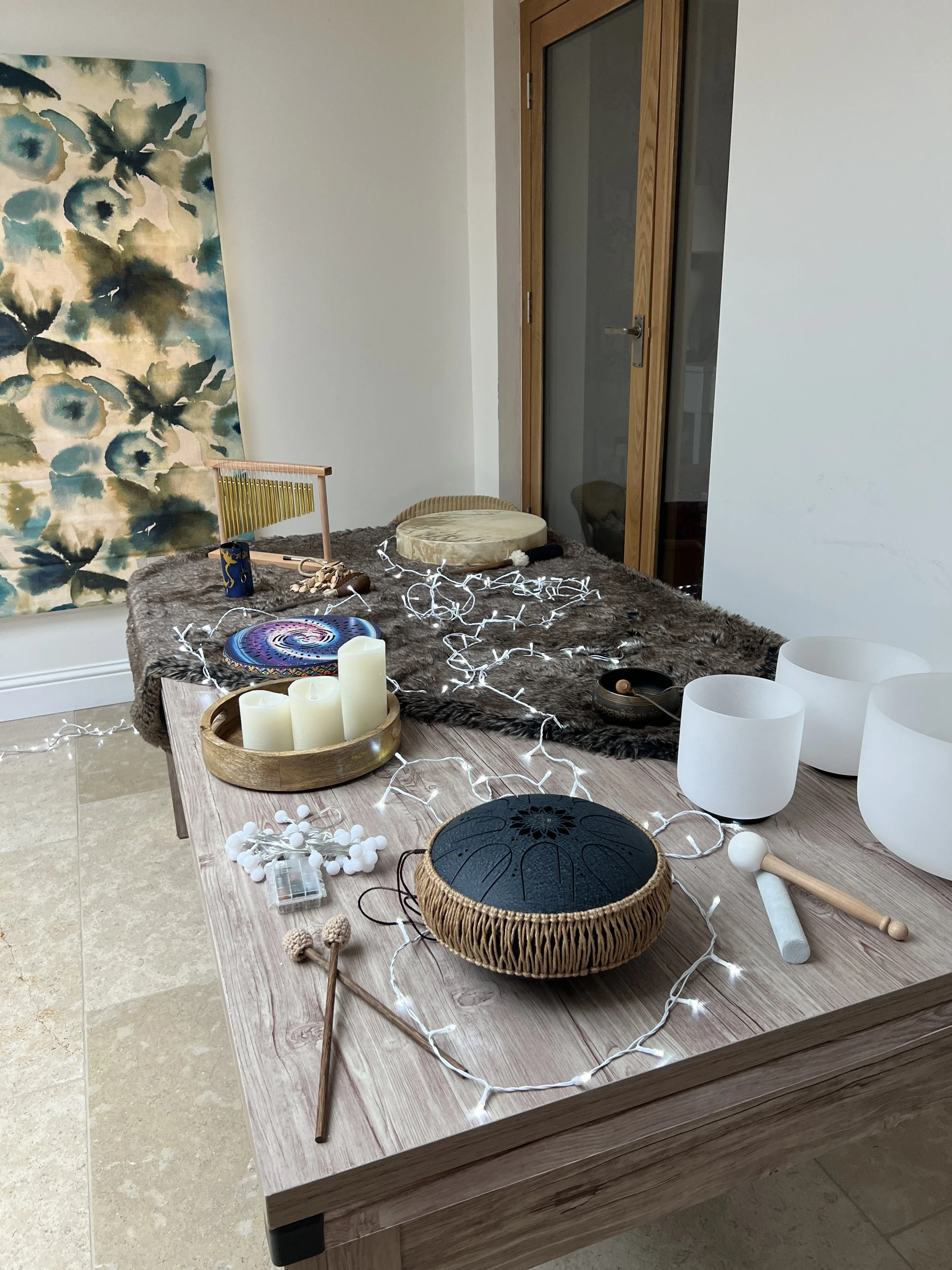 A wooden table decorated with candles, a black singing bowl with a woven cover, white bowls, fairy lights, and various percussion instruments, with a fur table runner, and art on the wall in a room.