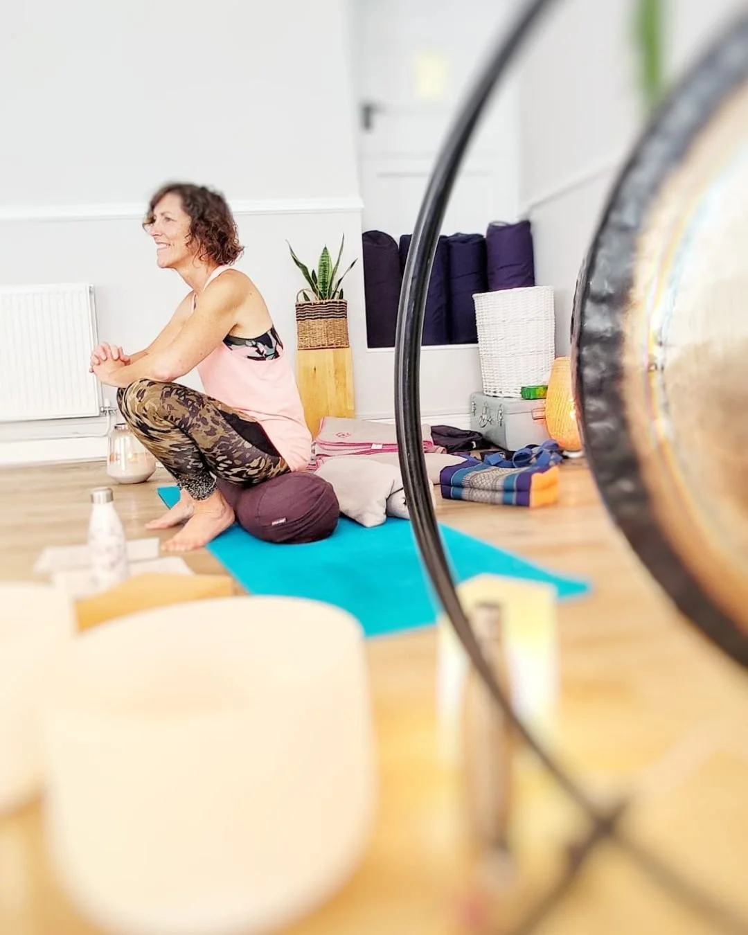 A woman practicing yoga in a bright room, sitting on a cushion with a yoga mat and various yoga props around her, viewed through a camera lens.