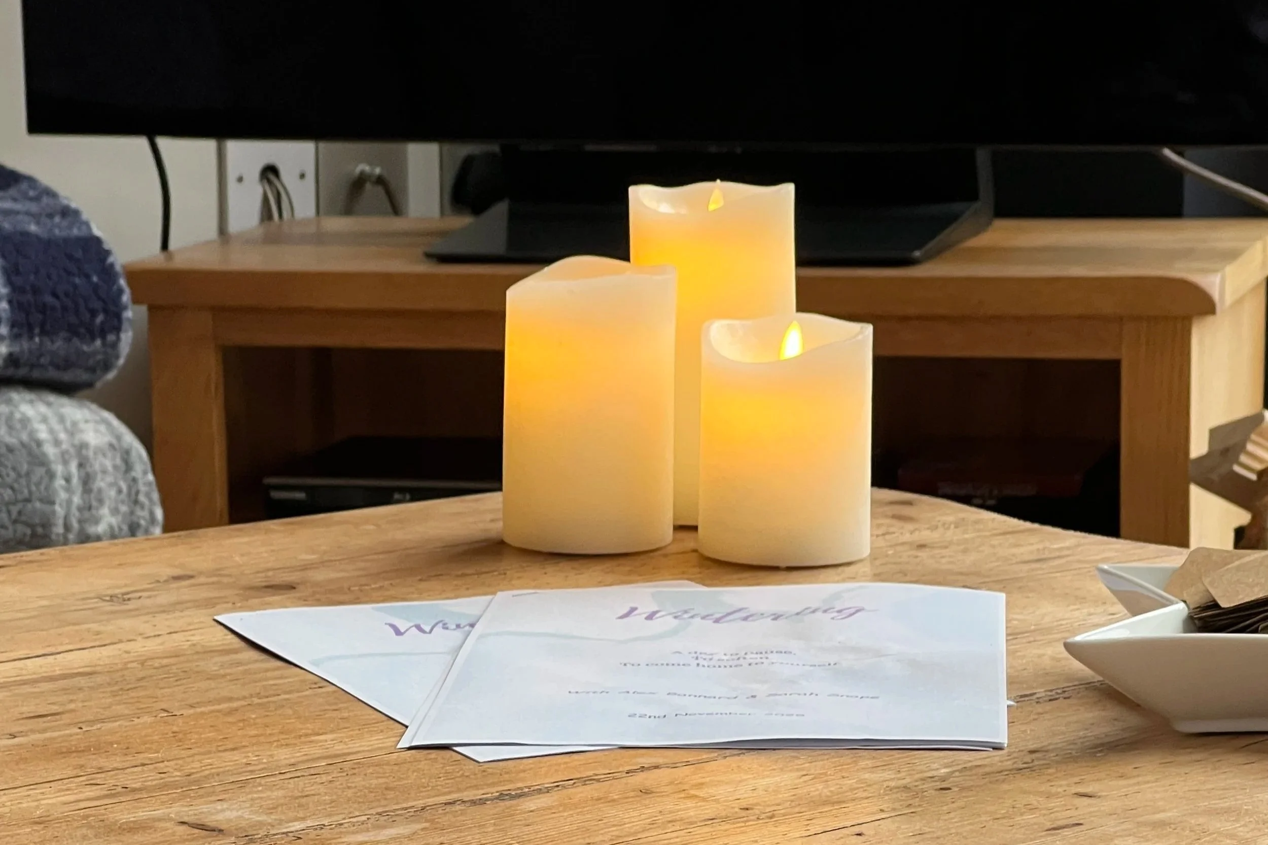 Wooden table with three lit candle fake candles, welcoming signs, and a small white dish with utensils, with a TV and wooden furniture in the background.