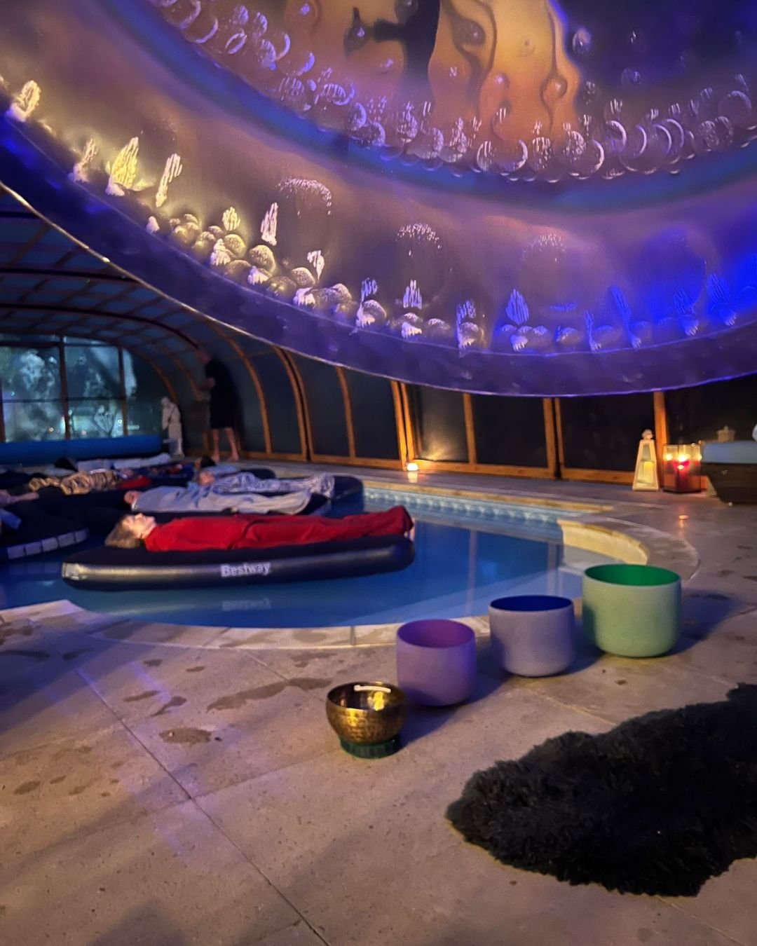 Indoor pool area with people lying on float beds, and colourful musical instruments near the pool for a floating sound bath experience.
