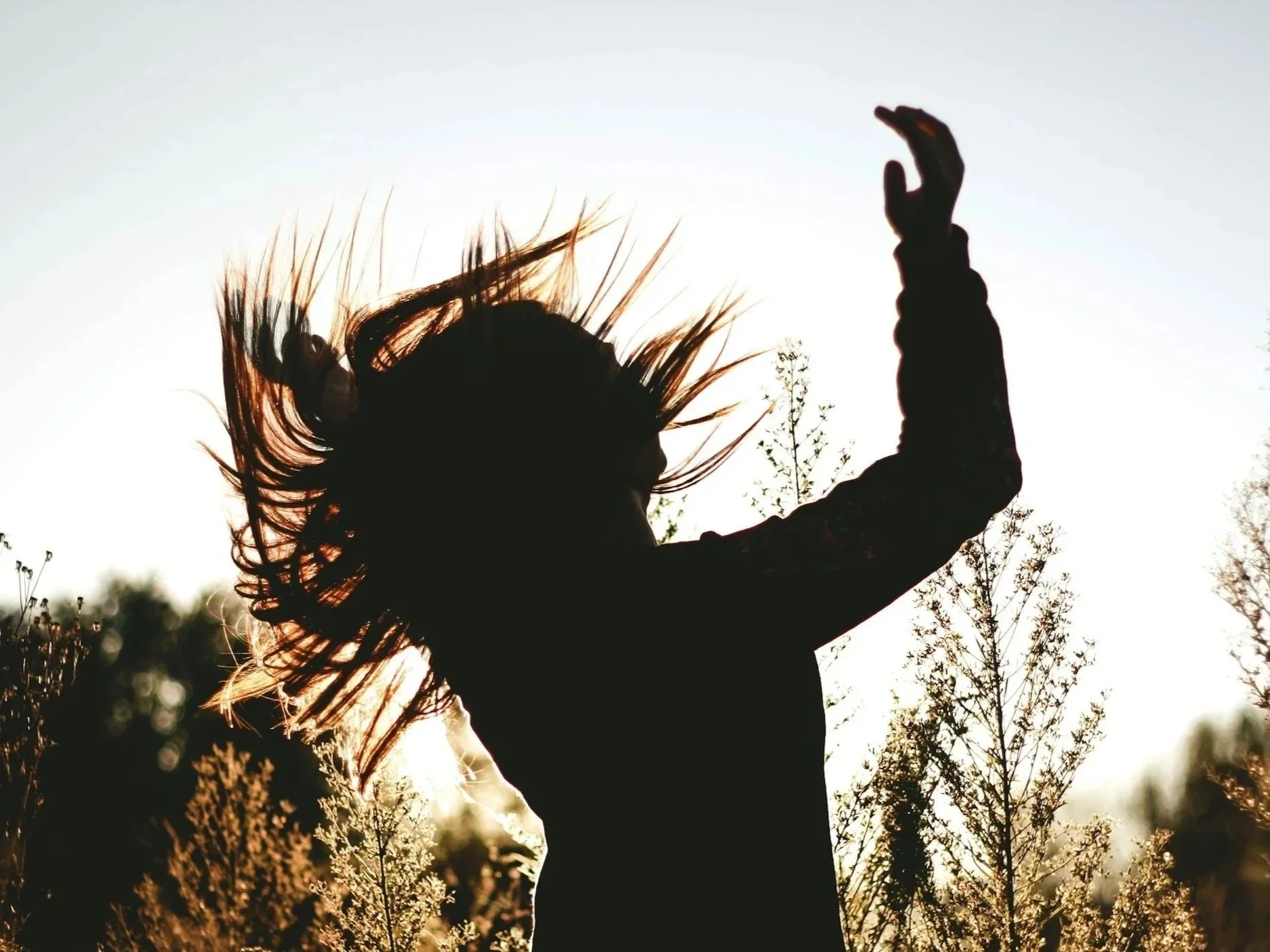 Silhouette of a woman with her hair and dress flowing, raising her right arm while standing outdoors at sunset or sunrise.