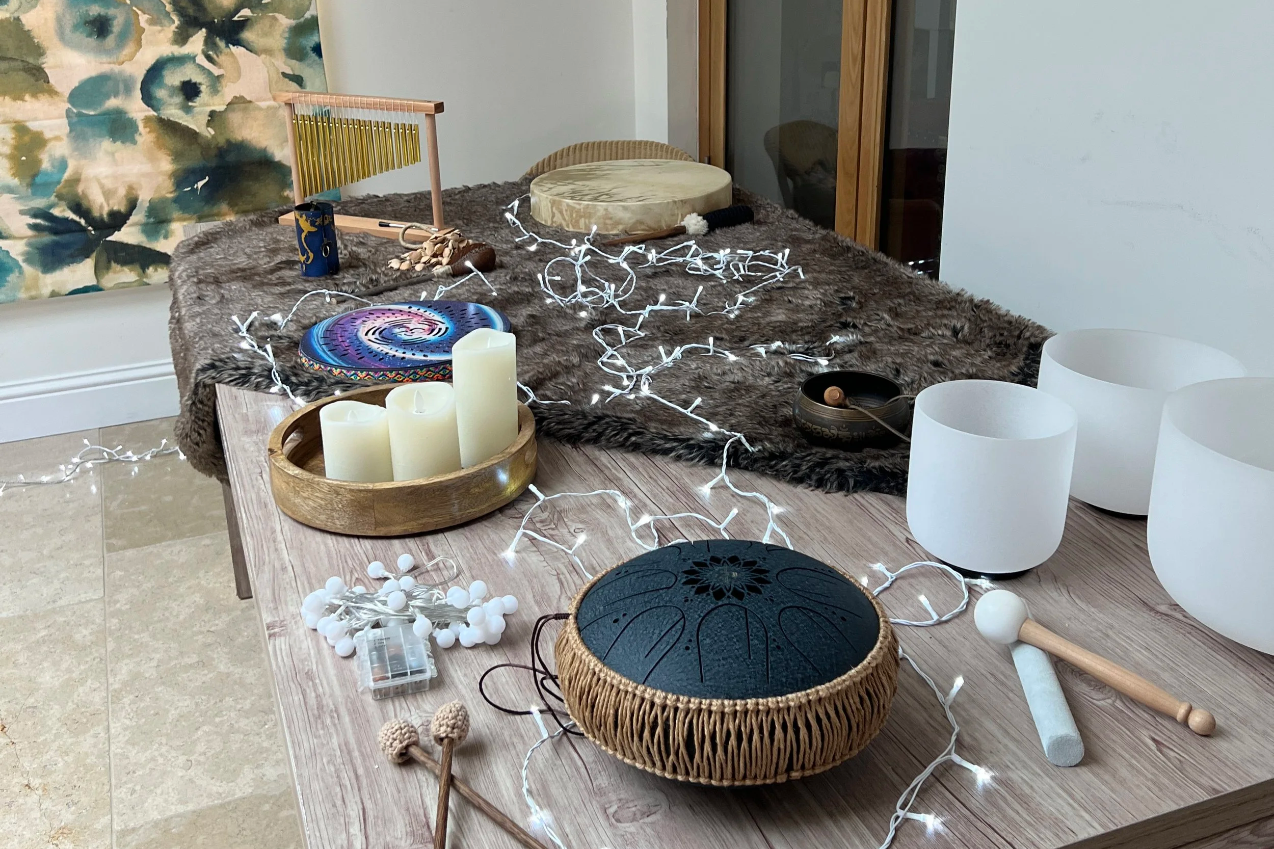 Array of crystal singing bowls, a faux fur cloth, candles, and musical instruments on a dining table decorated with string lights.