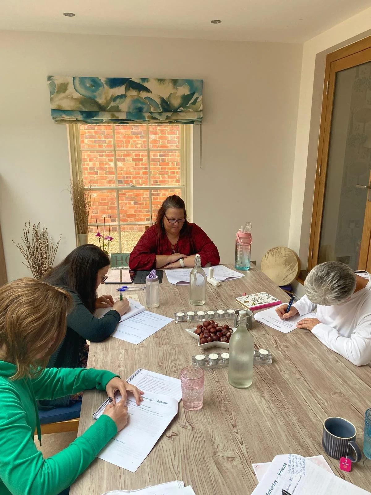 Four women sit around a wooden table in a bright room with a window, engaged in note-taking and discussion.