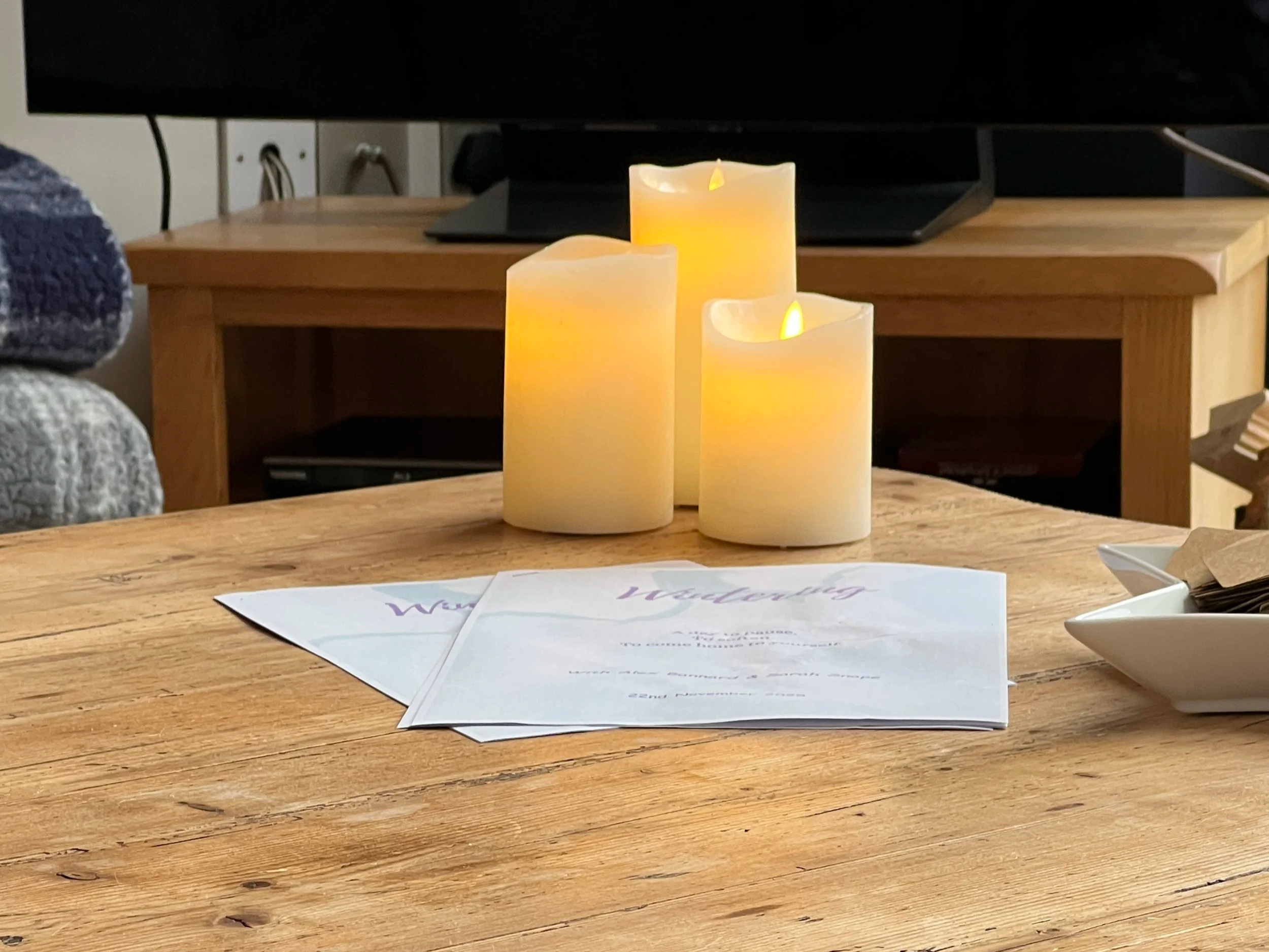 Three flameless candles on a wooden table with workbooks and a white dish.