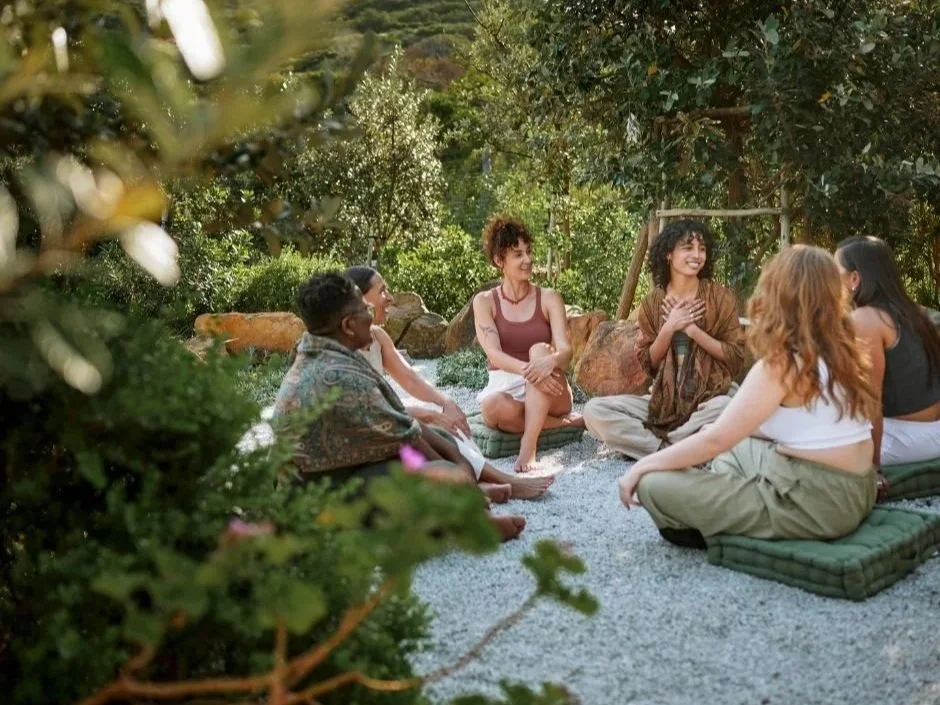 A group of five women sitting on cushions outdoors, engaging in a circle conversation amidst greenery and trees.