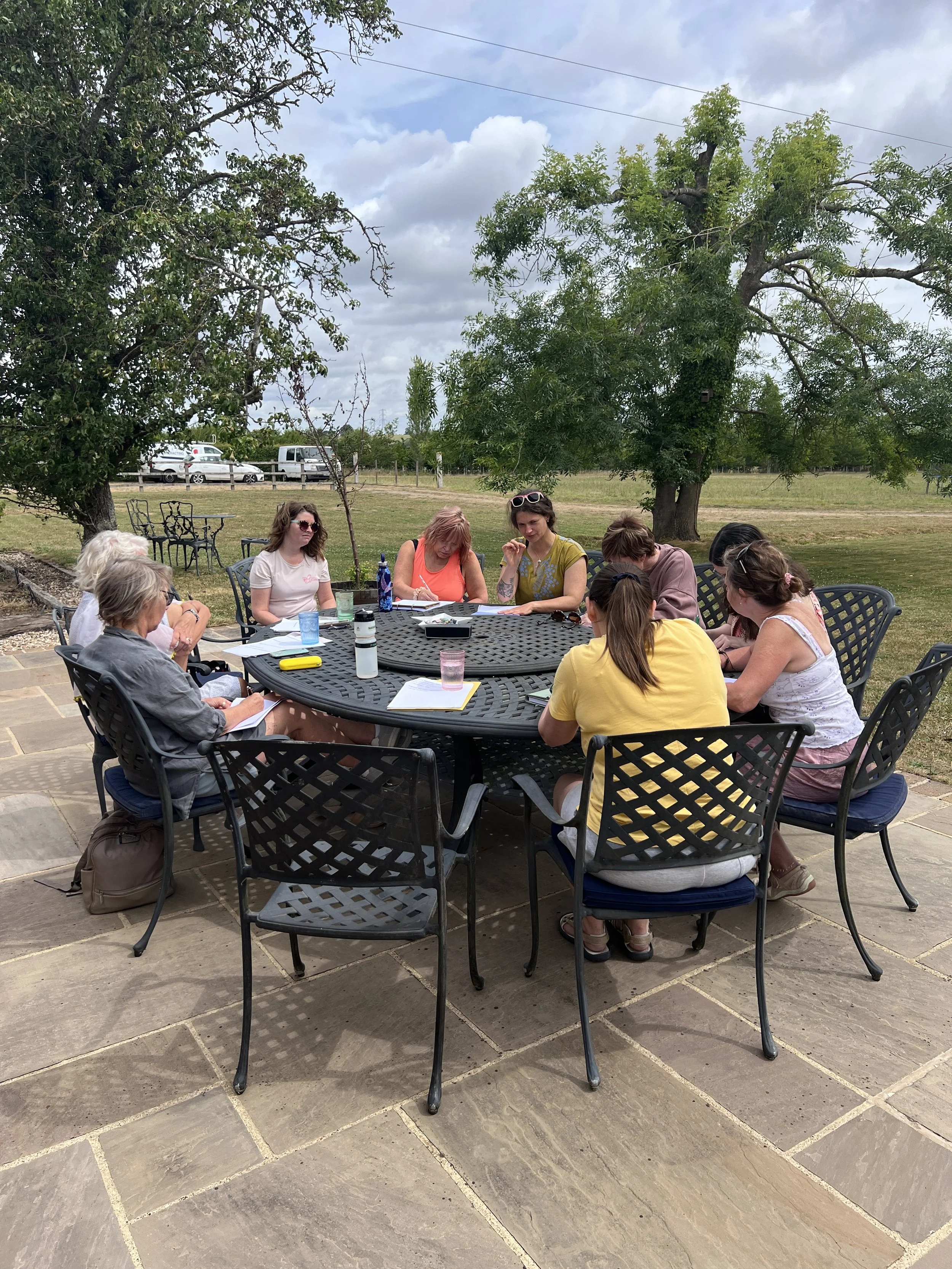 A group of people sitting around a round outdoor table on a patio, engaged in a retreat reflection. There are papers, notebooks, and water bottles on the table. Behind them is a grassy field with trees and a few parked cars under a partly cloudy sky.