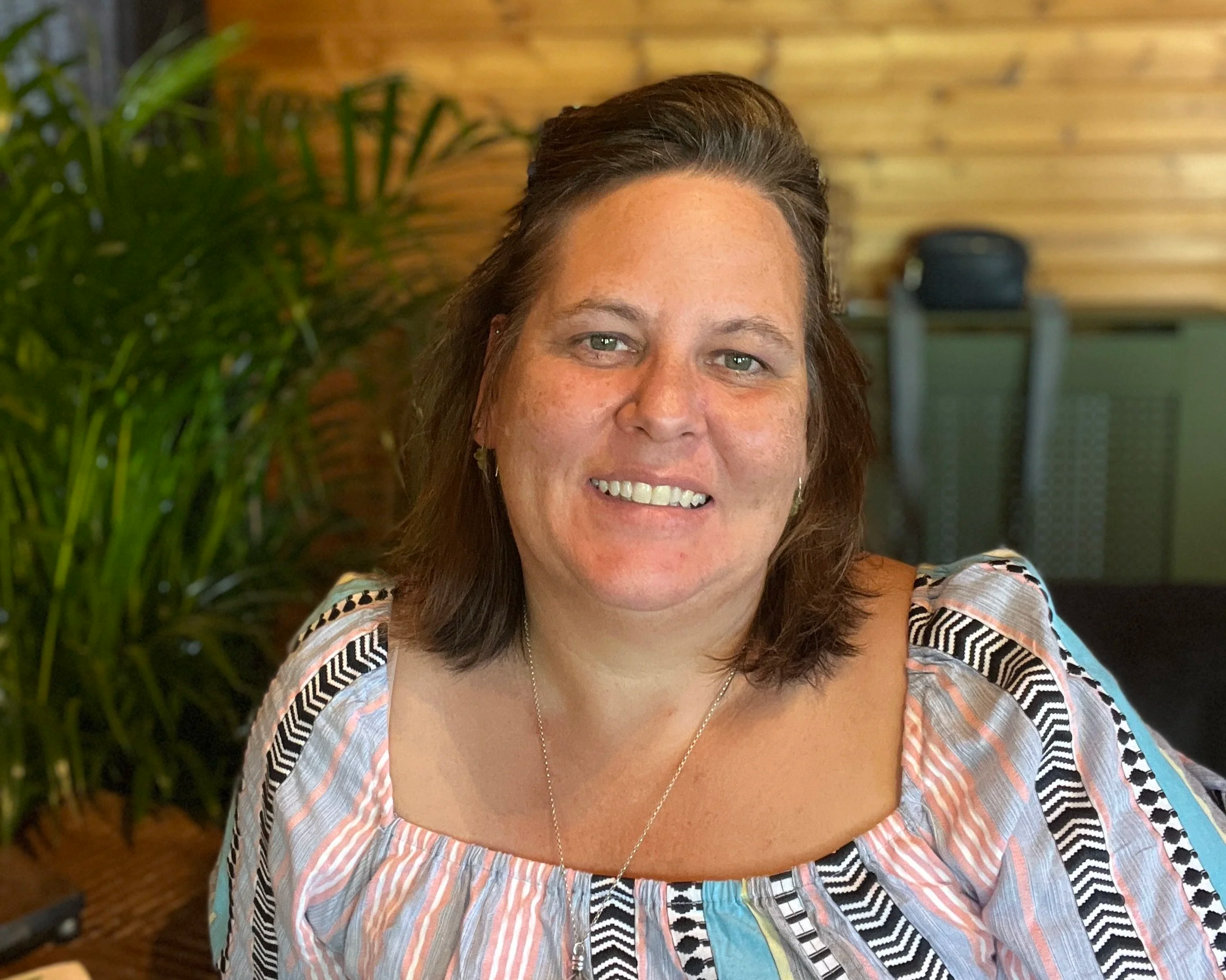 A woman with shoulder-length brown hair and light skin smiling at the camera, wearing a colorful patterned blouse, with a potted plant and a wood-paneled wall in the background.