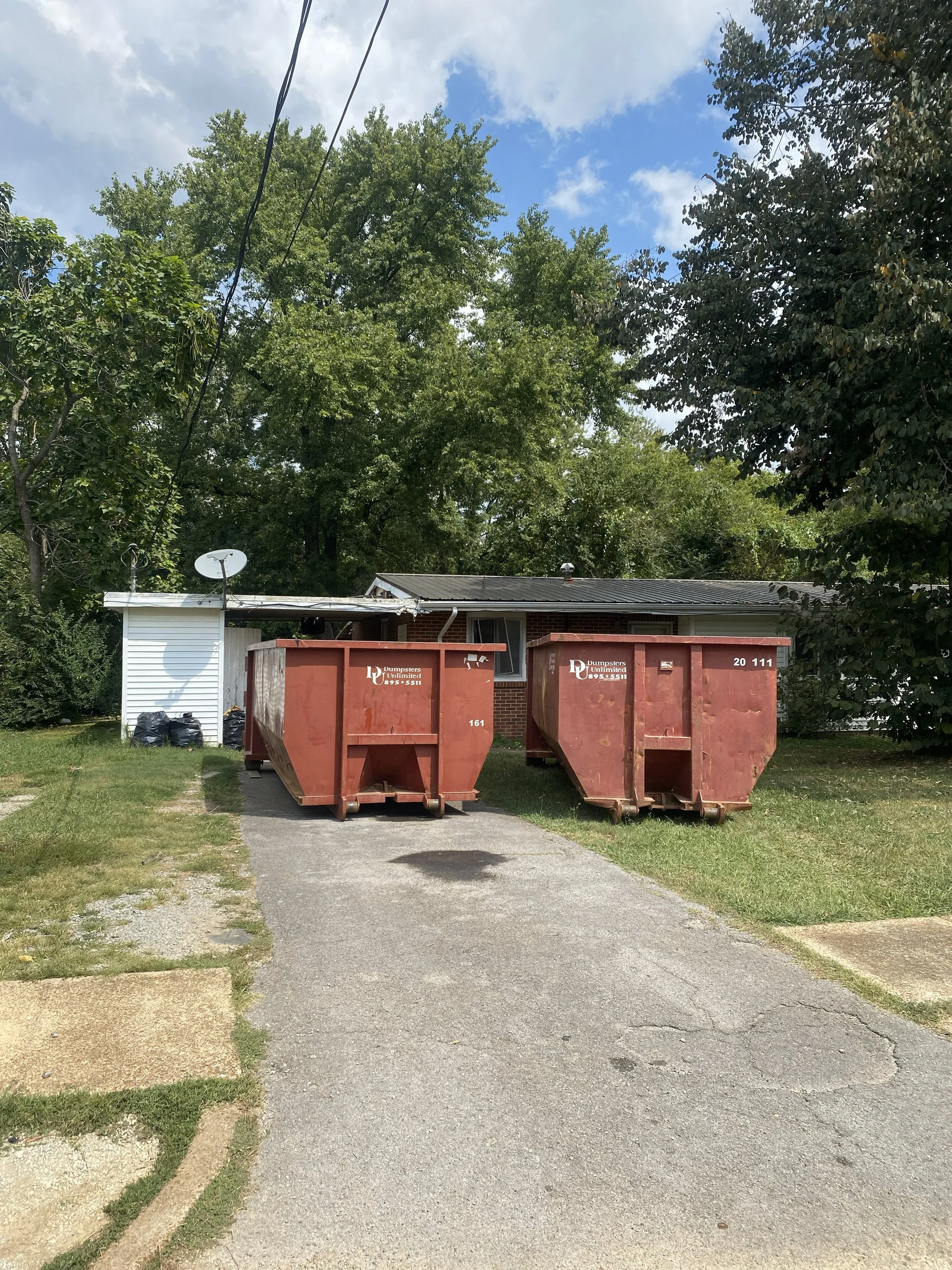 Two large red dumpsters in a residential driveway in front of a single-family house, with green trees and a partly cloudy blue sky in the background.