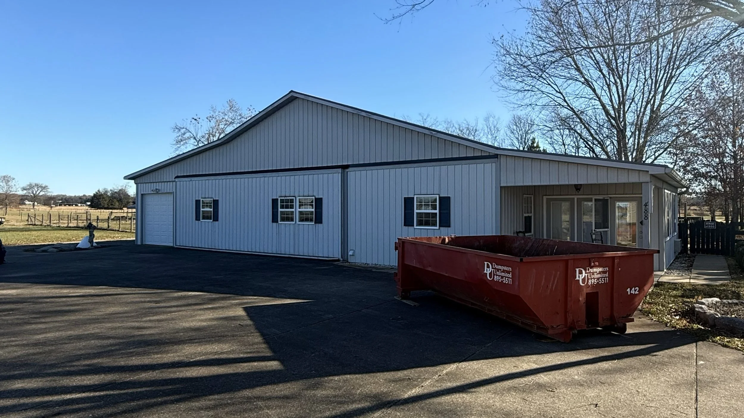 A white metal building with black shutters and a large sliding door, located on a paved lot, with a red dumpster labeled Dumpsters Unlimited in front and leafless trees in the background.