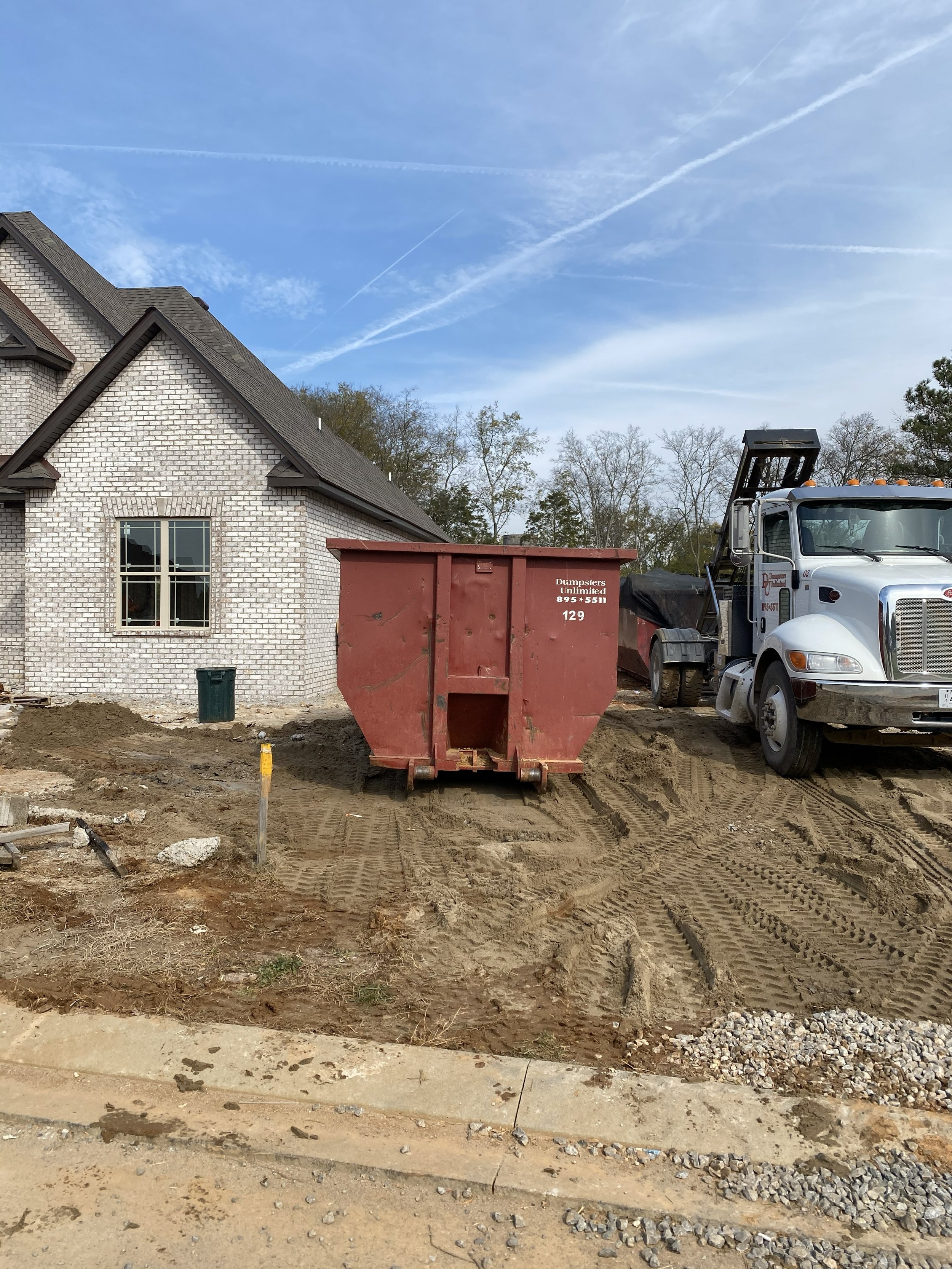 Construction site with a house in the background, a red dumpster, a large white truck, and dirt with tire tracks.