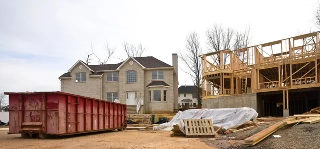 Construction site with a house under construction, wooden framework for another building, and a large red dumpster in the foreground.