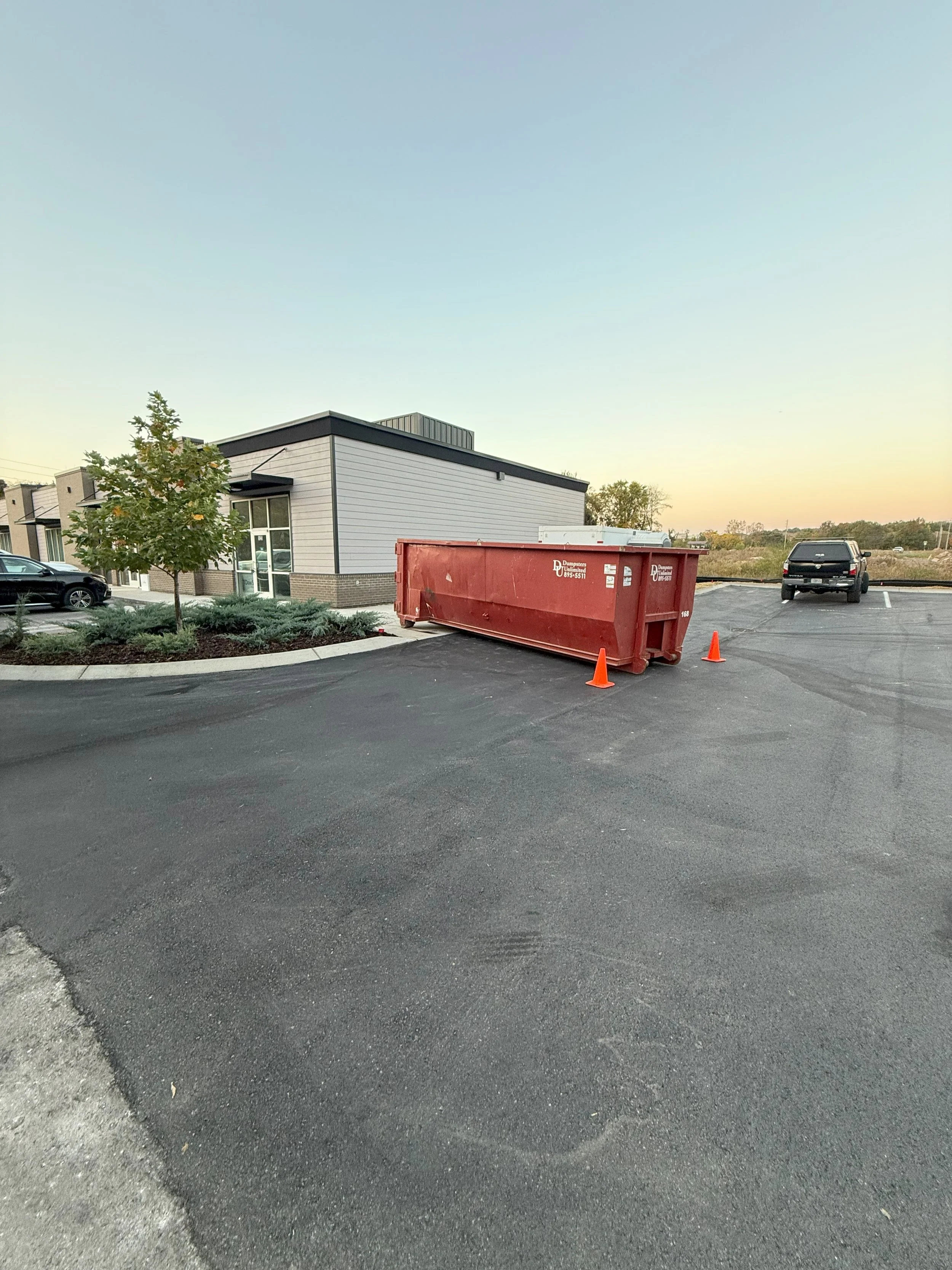 Empty parking lot with a large red dumpster and two orange traffic cones, modern building and clear sky in the background.