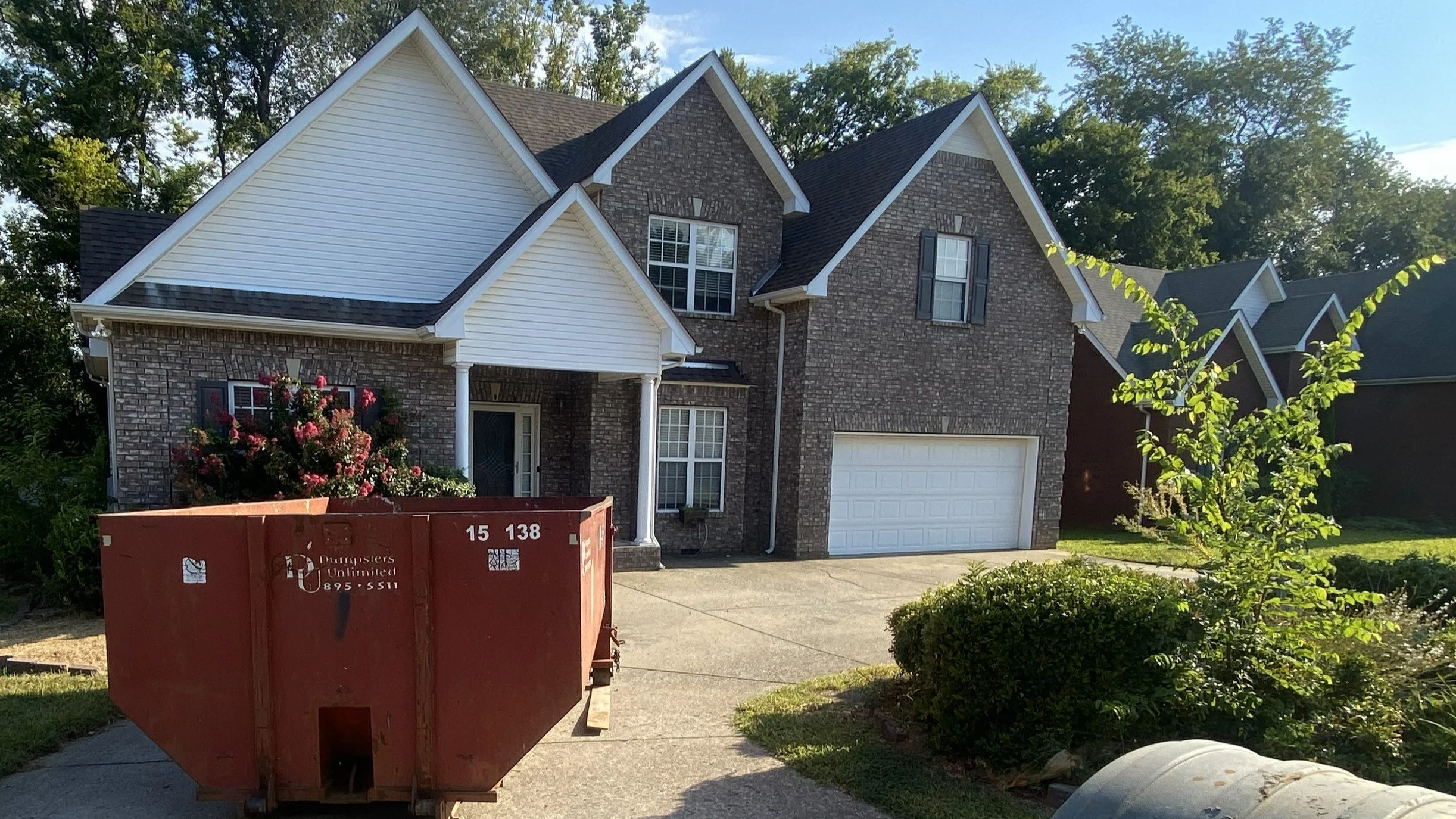 A two-story house with brick and white siding exterior, a white garage door, and a front porch with white columns. There is a red dumpster in the driveway and some bushes and small trees in the yard. The sky is clear with some clouds, and there are o