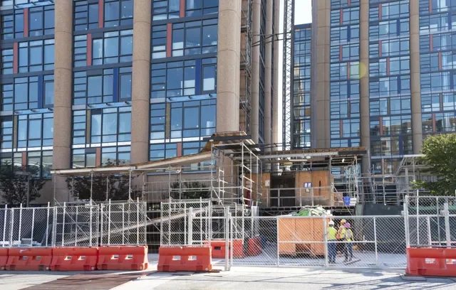 Construction site in front of a modern glass office building, with orange safety barriers and workers wearing helmets and high-visibility vests.