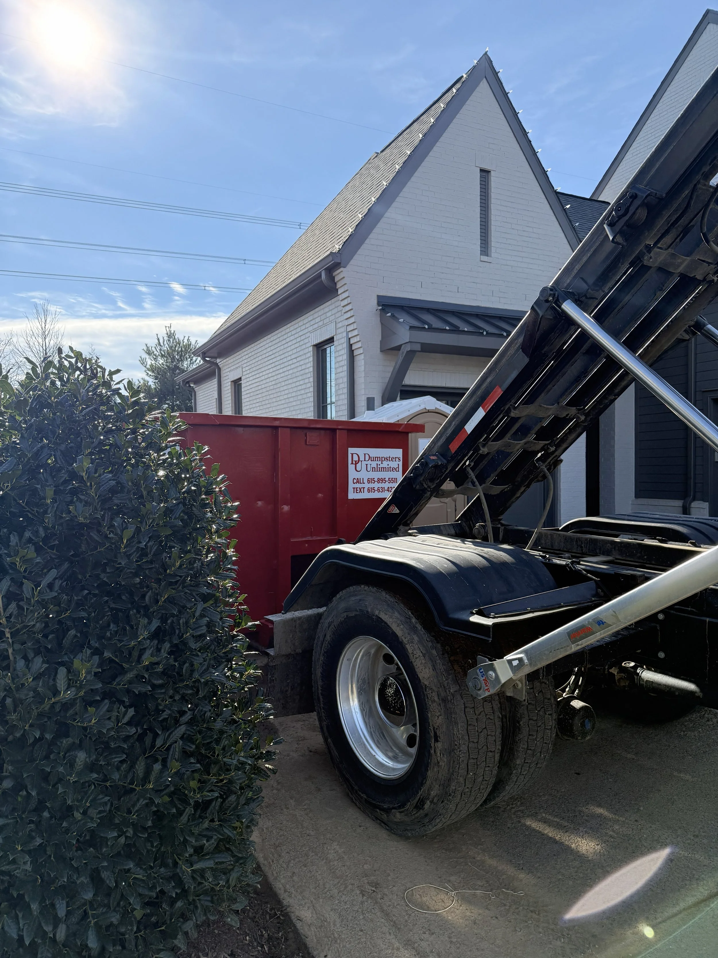 A house with white brick exterior and steep roof, with a truck and a red dumpster labeled 'Dumpster Unlimited' in the driveway during daytime