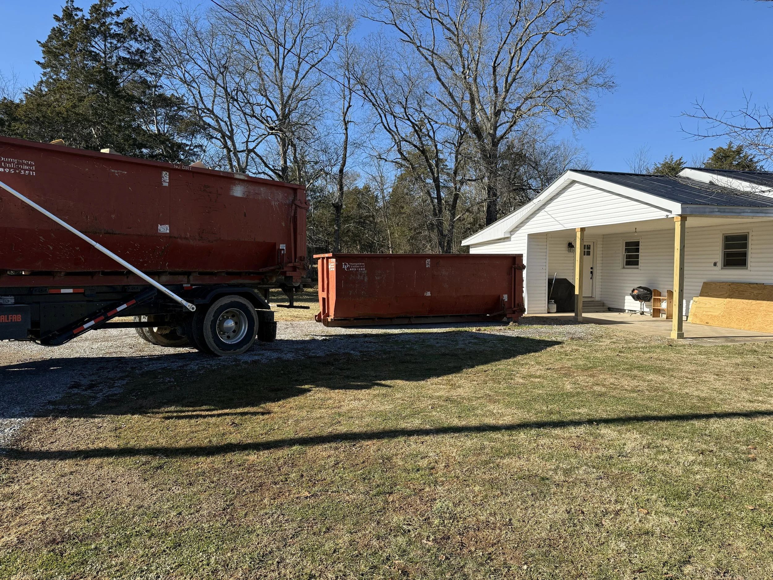 A white house with a black roof and a small covered porch, with a black grill and a wooden bench, is seen in a yard with a gravel driveway. A large red dumpster and a smaller red dumpster are parked nearby on the grass.