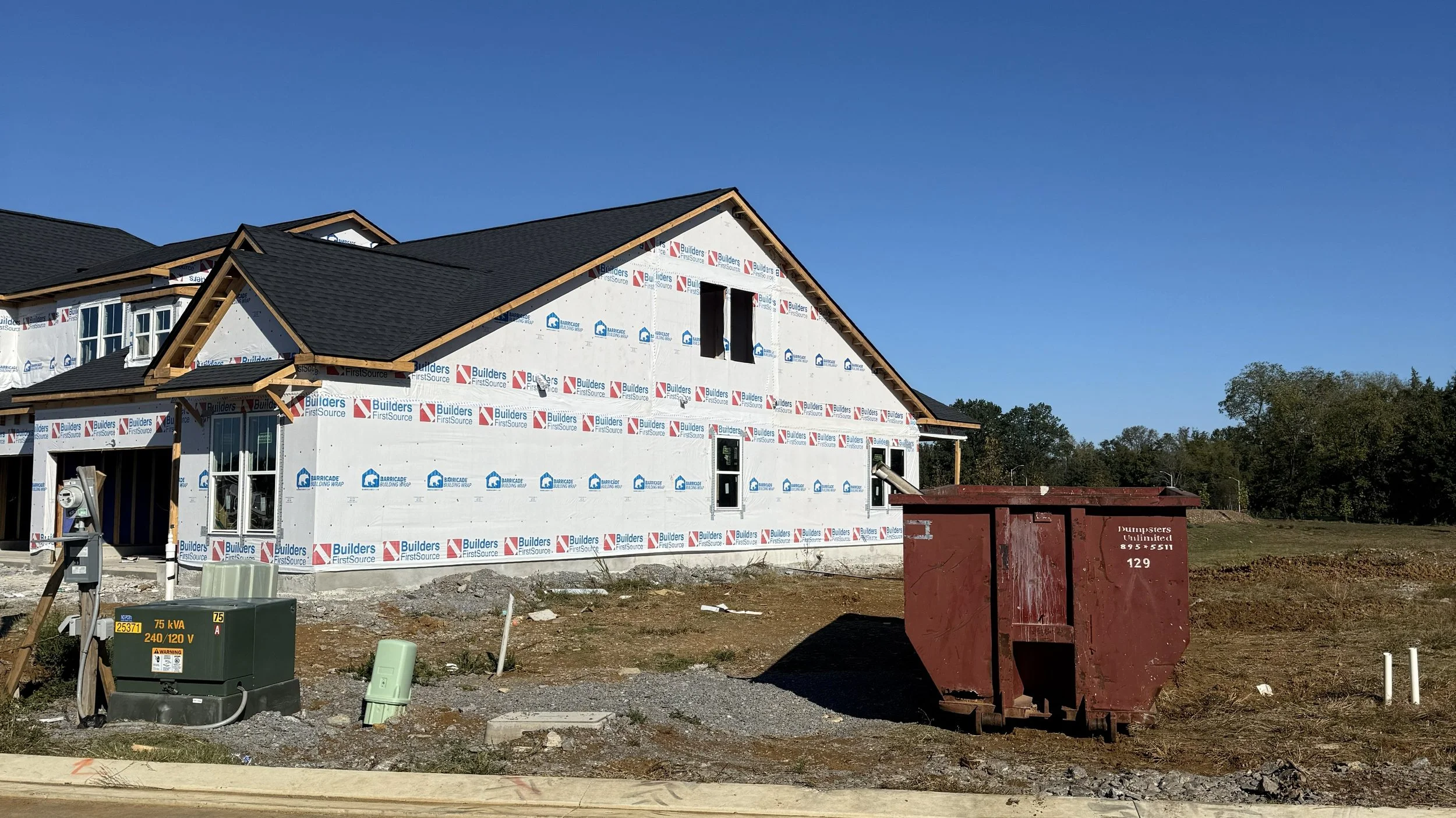A house under construction with black shingles roofing and partially completed exterior walls, on a clear day with blue sky and trees in the background. A large red dumpster is in the foreground, along with utility equipment and a utility box.