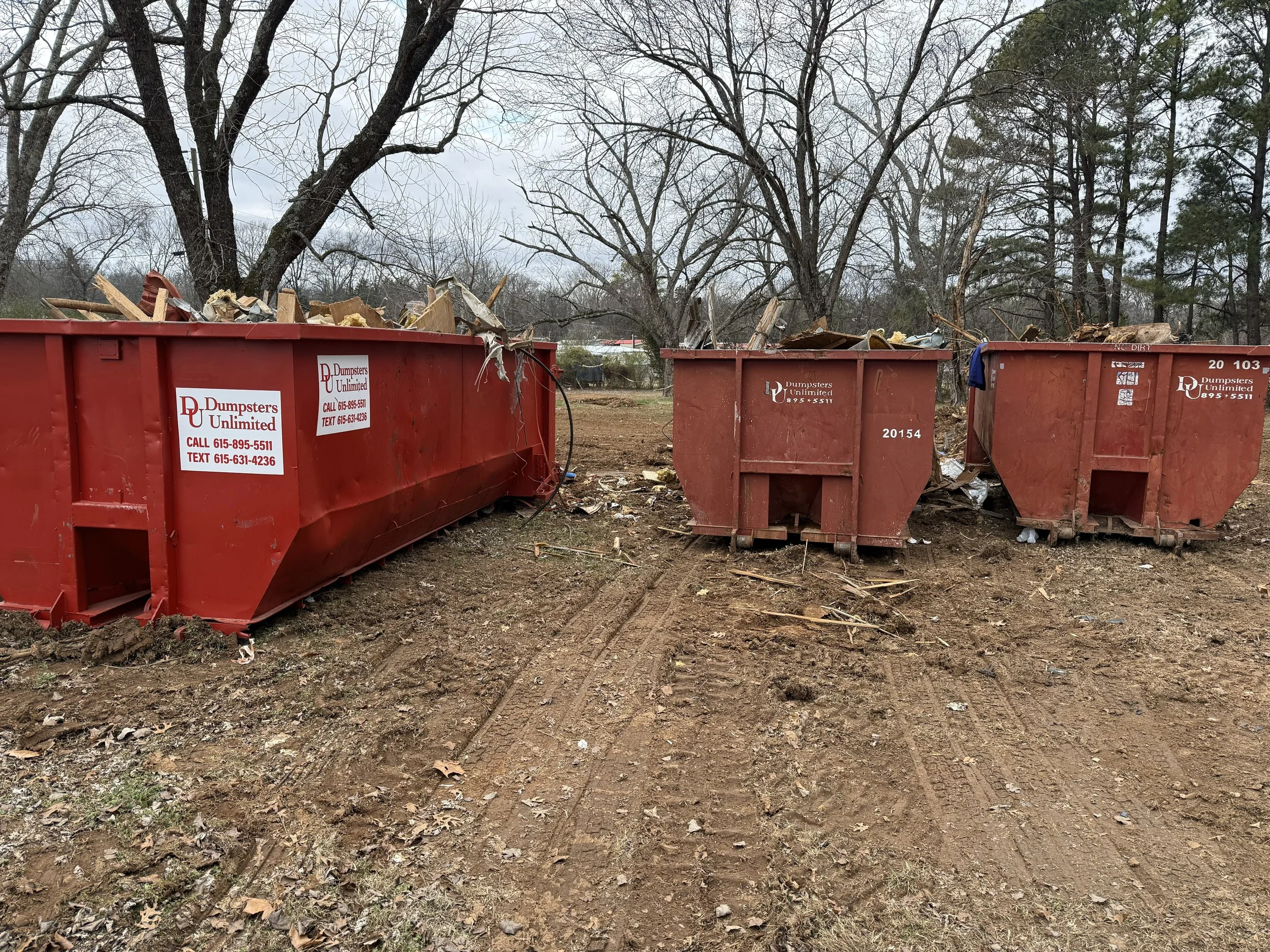 Three large red dumpsters filled with construction debris, placed outdoors on dirt ground with tire tracks, surrounded by leafless trees under a cloudy sky.