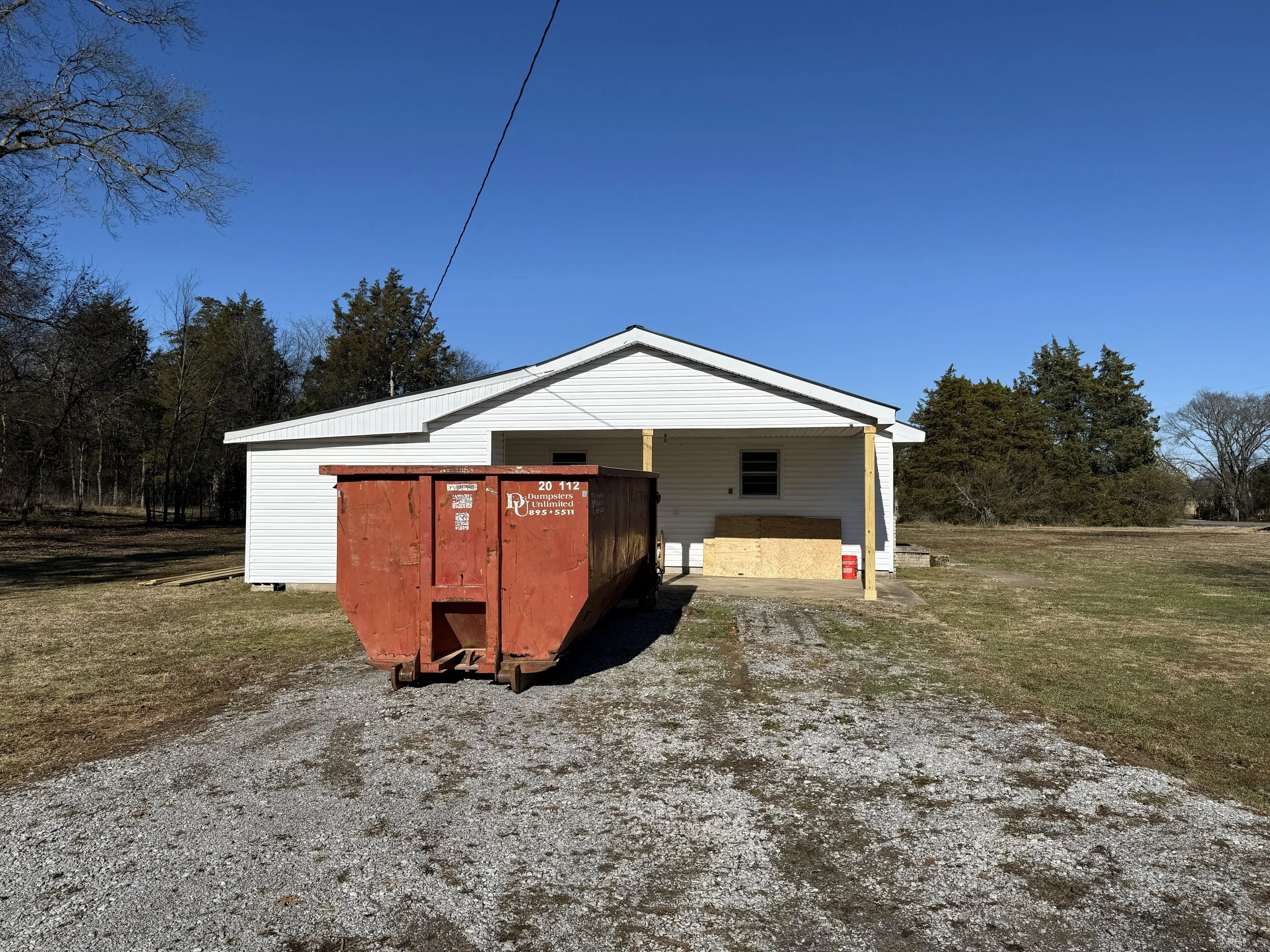 A white house with a small window, a wooden post on the front porch, a red dumpster, and an open yard with trees surrounding the property.