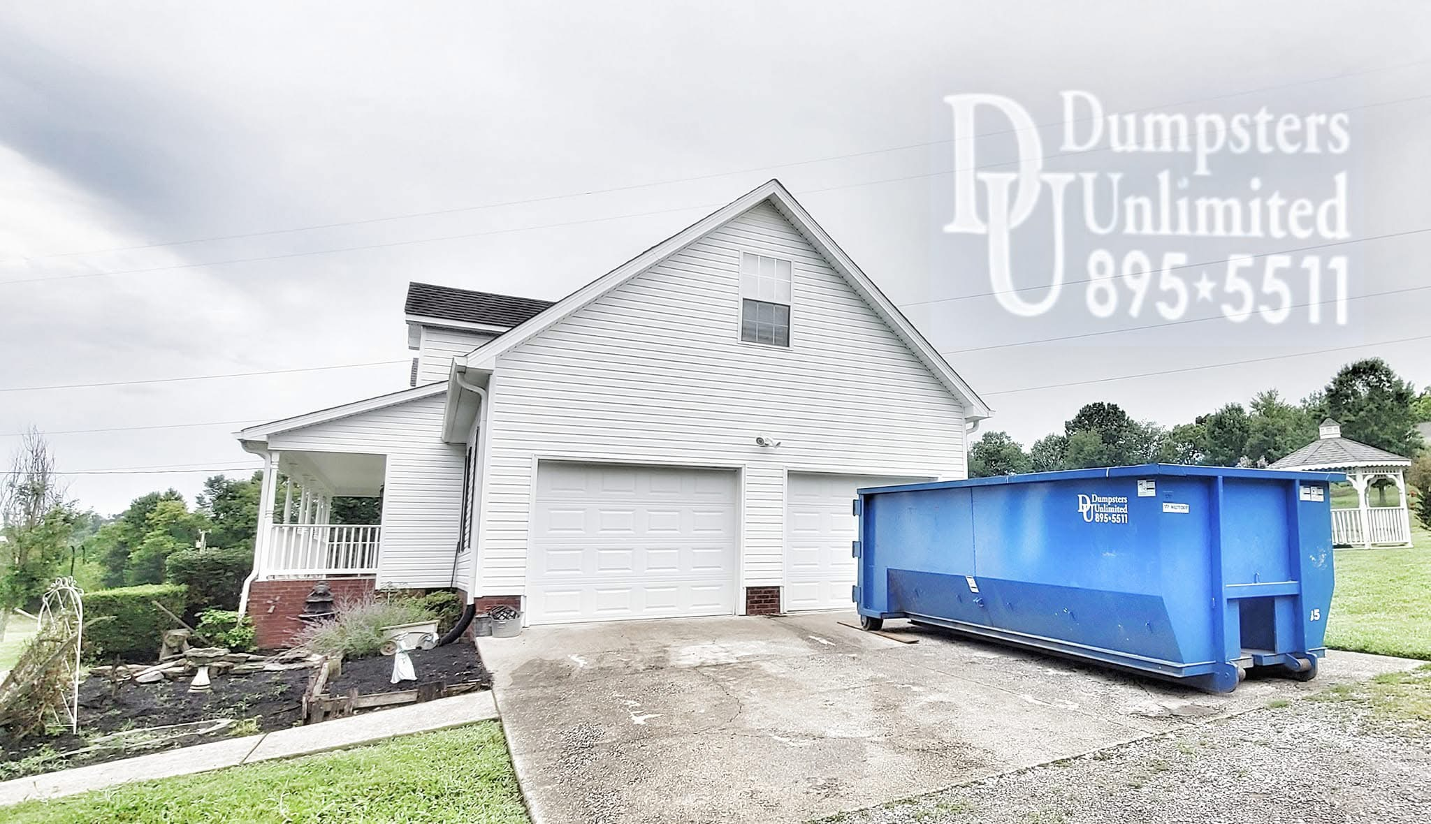 A white house with a two-car garage and a large blue dumpster in the driveway. The house has a porch on the left side and a small window above the garage. There is a garden bed with plants and a white arch on the left, and a grassy area with trees in the background under an overcast sky.