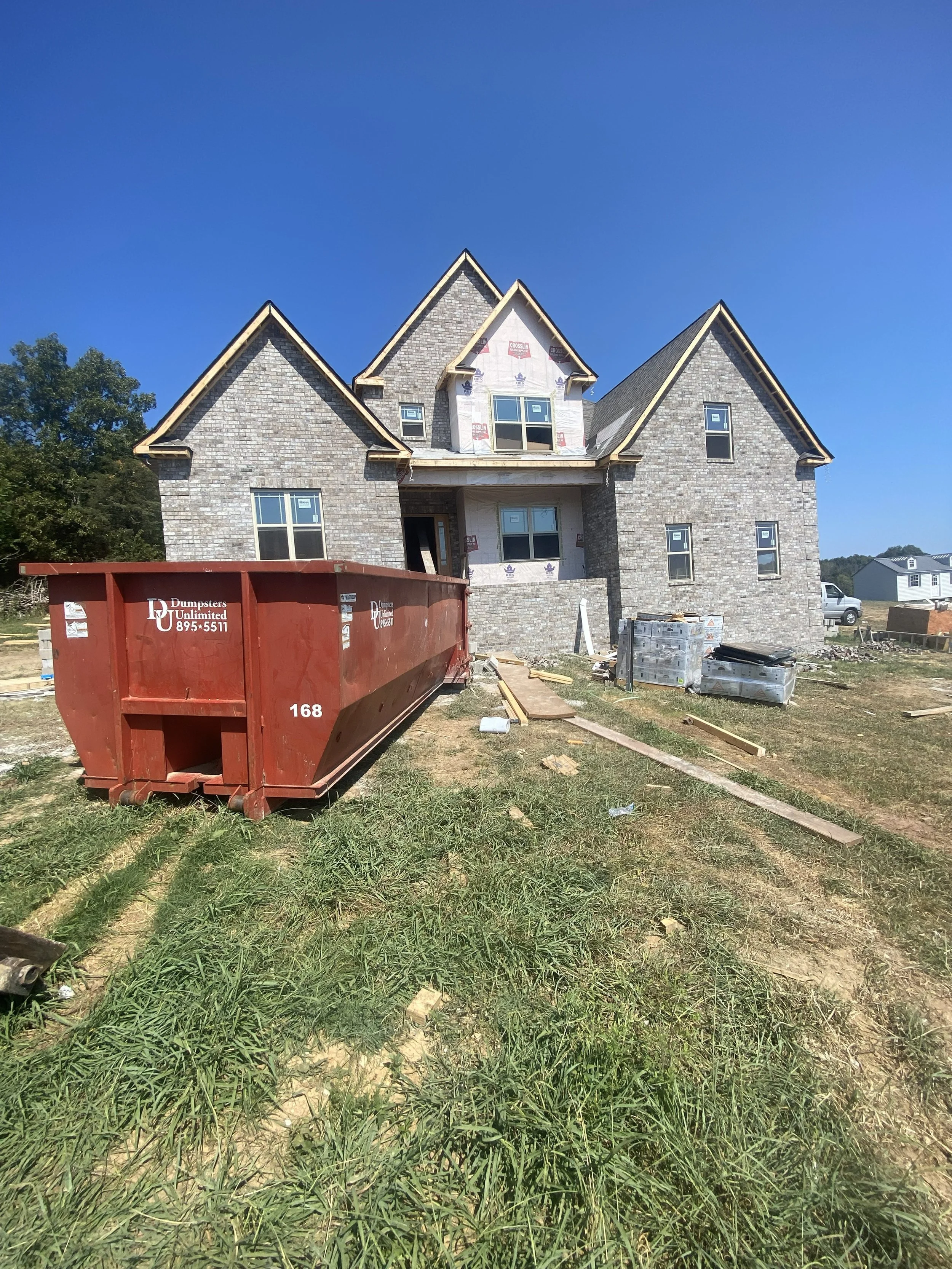 A house under construction with brick exterior, construction materials, and a large red dumpster in the front yard on a clear, sunny day.