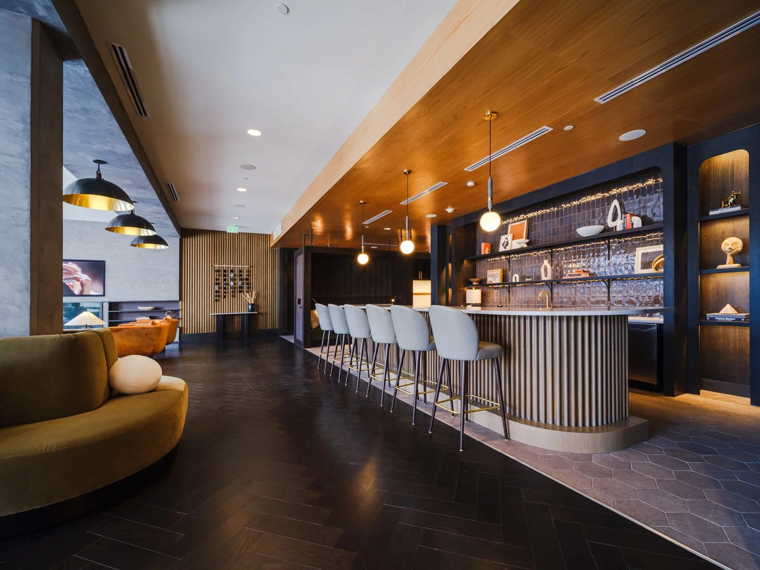 Modern bar with a curved bar counter, white barstools, warm wooden ceiling, black shelving with decorative items, and dark wooden flooring.