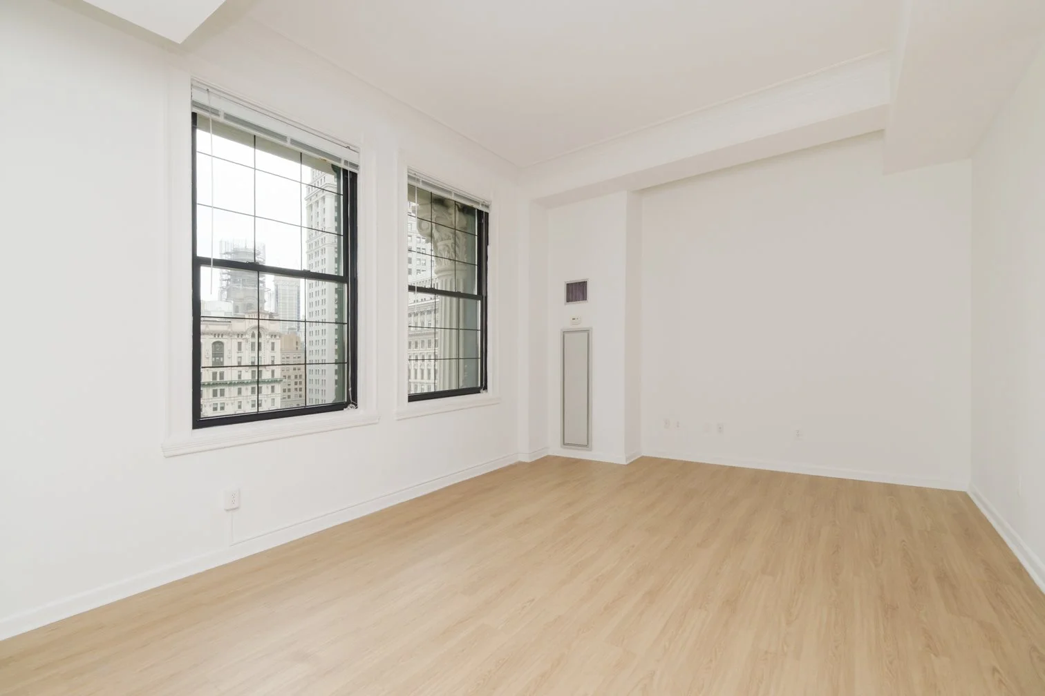 Empty room with hardwood floors, white walls, and large windows showing a cityscape with tall buildings.