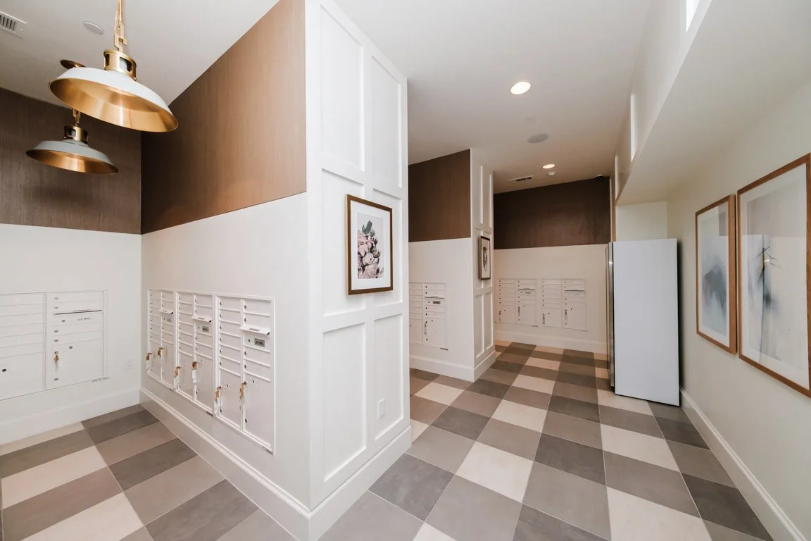 An hallway with mailboxes on the wall, framed art, and a refrigerator, with checkered tile flooring.