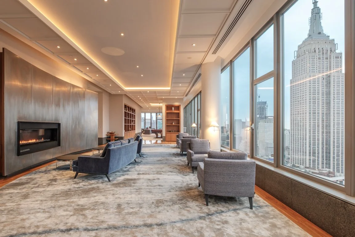 Luxurious high-rise apartment Residents Lounge with large windows showcasing city skyline including the Empire State Building, modern fireplace, gray sofas, armchairs, wooden shelves, and soft lighting.