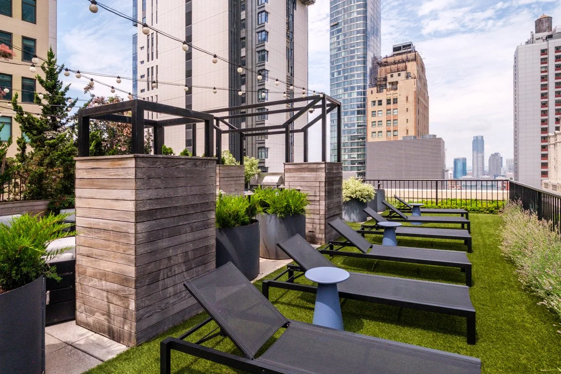 Rooftop terrace with black lounge chairs, small blue tables, planters with greenery, and city skyline in background