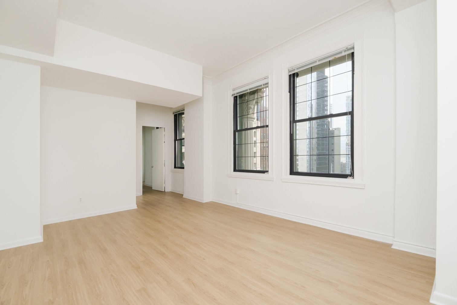 Empty room with white walls, light wood flooring, three large windows with black frames, and city buildings visible outside.