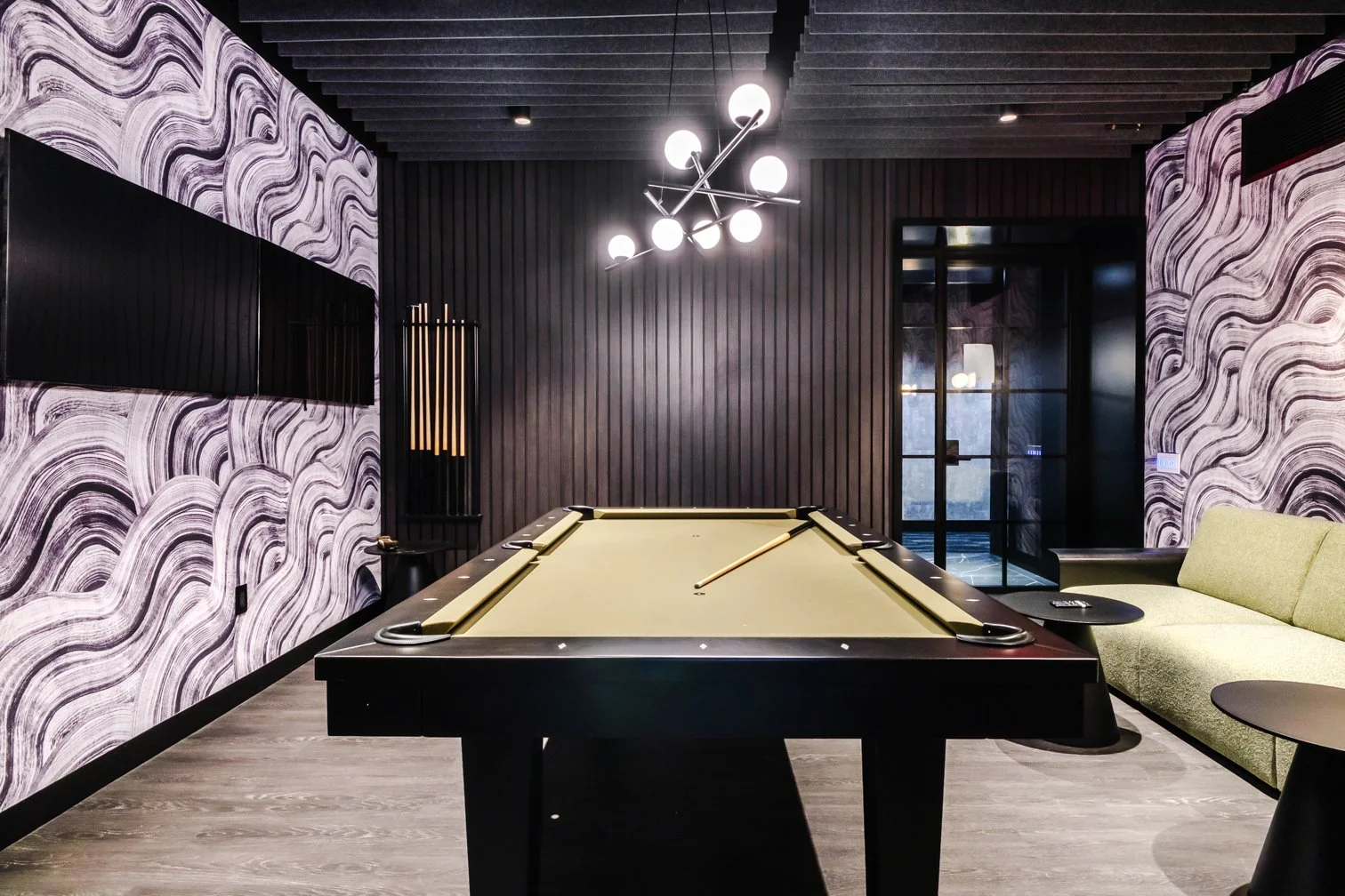 A modern game room with a black-felt pool table and a cue stick, green seating sofa, wall-mounted flat screens, and contemporary lighting fixtures.