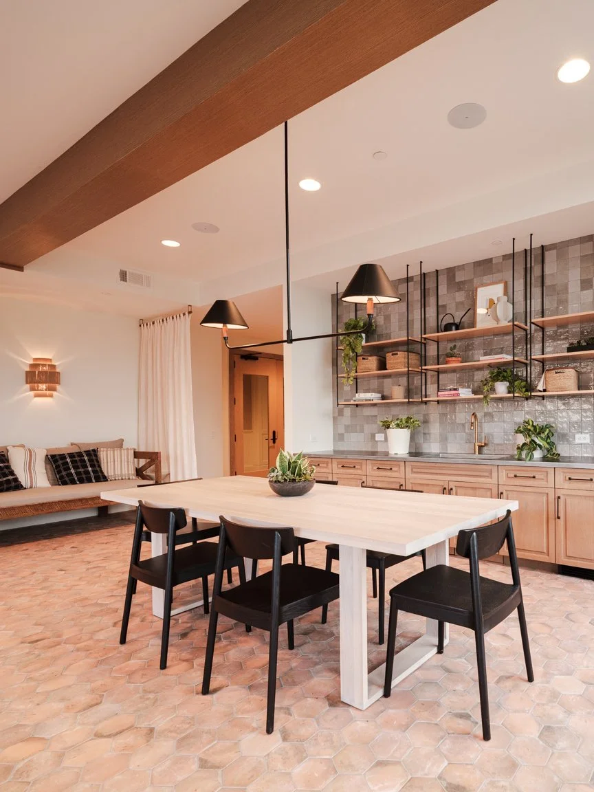 Modern kitchen and dining area with light wood table, black chairs, open shelving, plants, and pendant lights.
