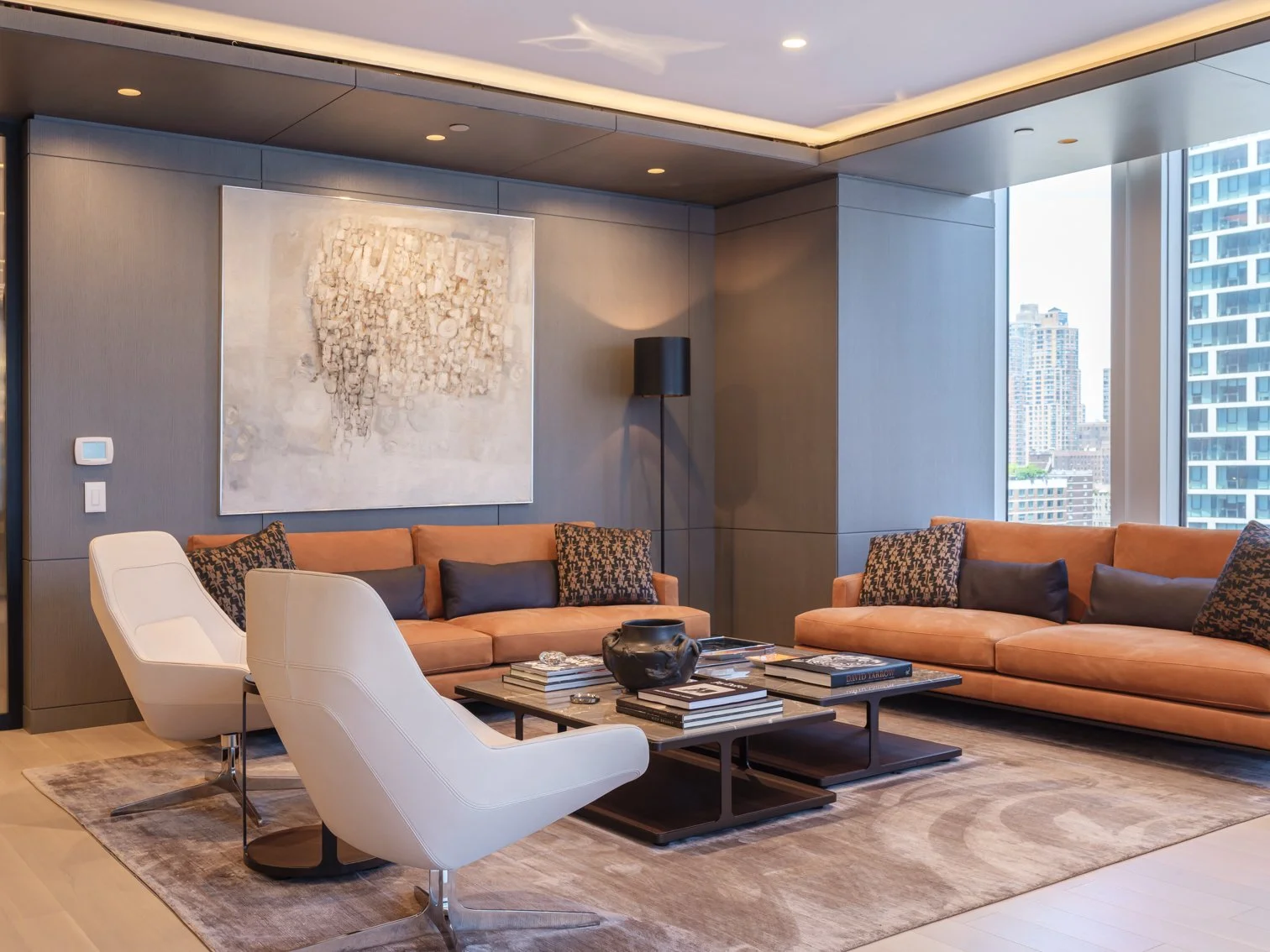 Modern executive office with two beige sofas, two white armchairs, a coffee table with books and a black vase, large window with city view, abstract wall art, and a gray wall with a black floor lamp.