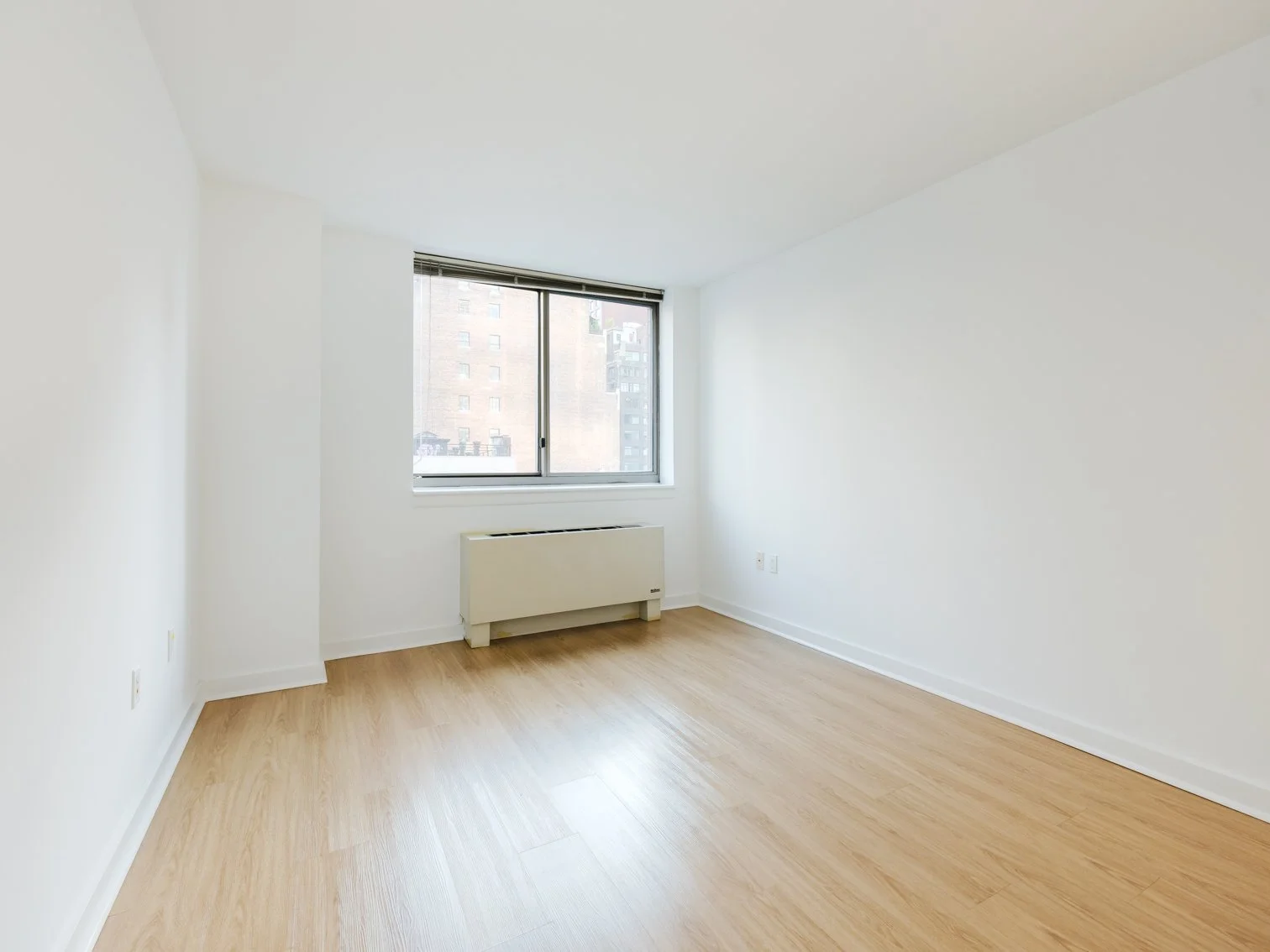 Empty room with white walls, wooden flooring, a large window, and a white heater below the window.