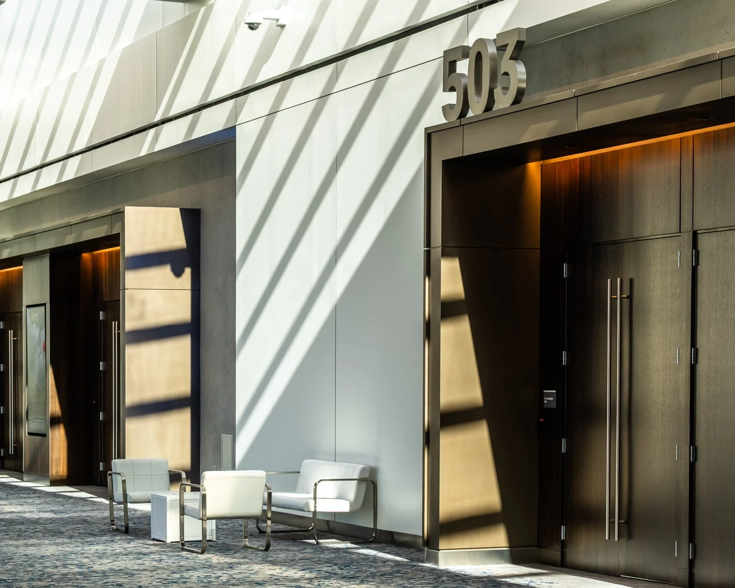 Modern building with number 503 above the entrance, featuring large glass and wood doors, chairs outside, and shadows cast by nearby structures.