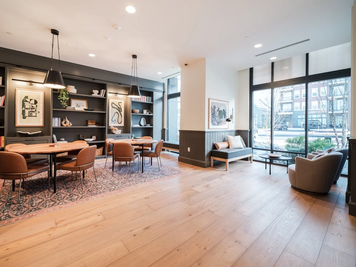 A modern, well-lit living or lounge area with large windows, comfortable seating, a rug, and a decorated wall with artwork and shelves.