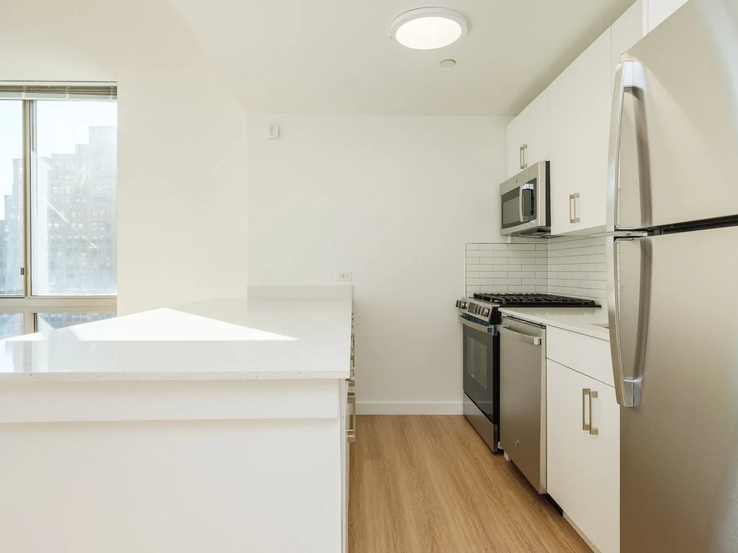 Modern kitchen with white cabinets, stainless steel refrigerator, microwave, stove, and white tile backsplash, illuminated by ceiling light.