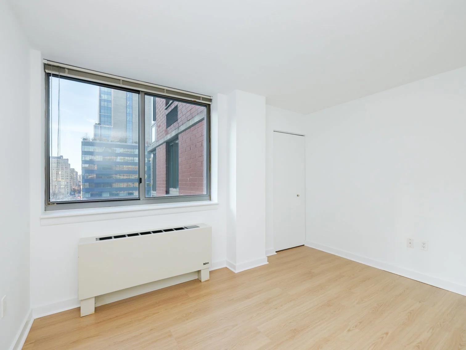 Empty room with white walls, light wood flooring, a large window showing tall buildings outside, a white radiator below the window, and a closed white door.
