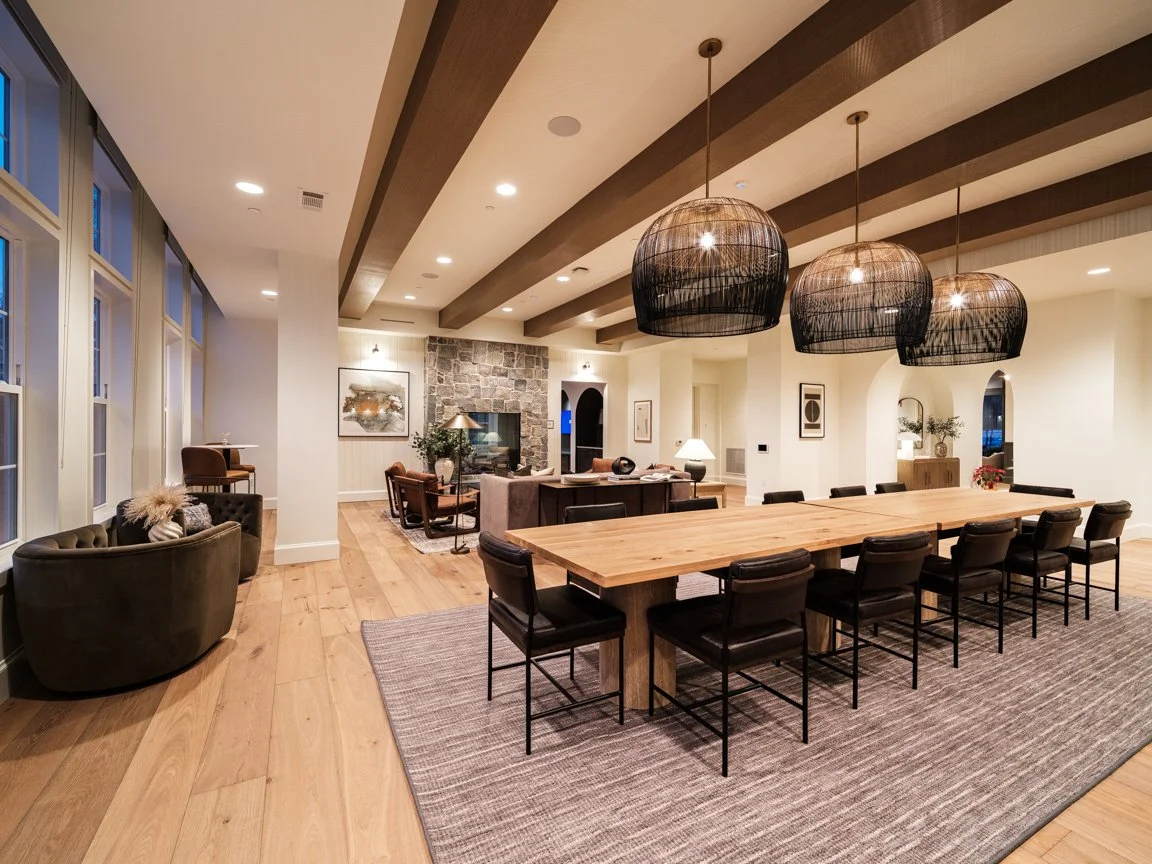 Spacious living and dining area with wooden flooring, large windows, a long wooden dining table, modern black chairs, a sitting area with armchairs, a stone fireplace, and contemporary artwork.