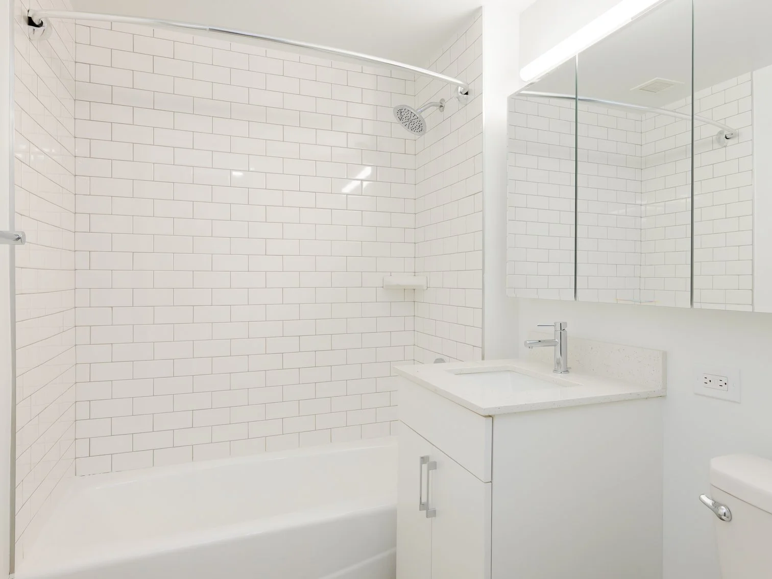 Minimalist bathroom with white subway-tile wall, bathtub, small vanity with sink, large mirror, and a toilet.