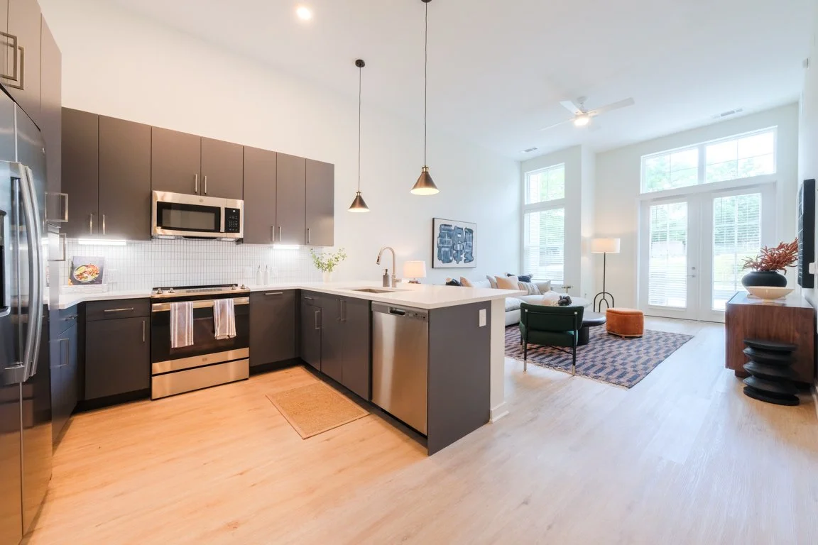 Open-concept kitchen and living room with large windows, modern gray cabinets, stainless steel appliances, and wooden flooring.